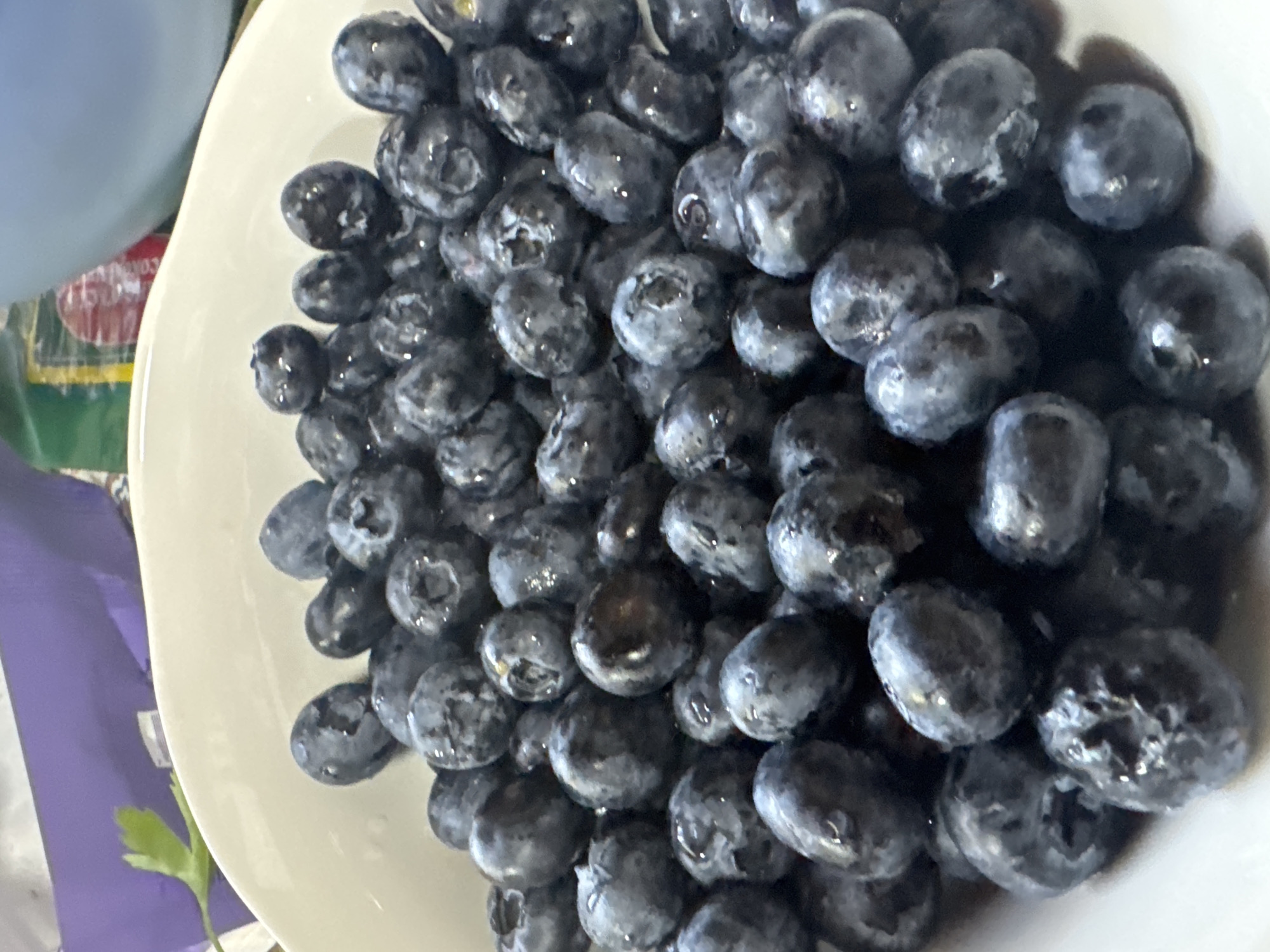 A white bowl is filled with a large quantity of blueberries. The berries are dark blue with a powdery bloom, and some appear to have water droplets on their surfaces. The bowl is sitting on a surface near a blue object, a purple object, and some greenery. There is no indication this photo was taken in Малая Рукавицкая, Russia.