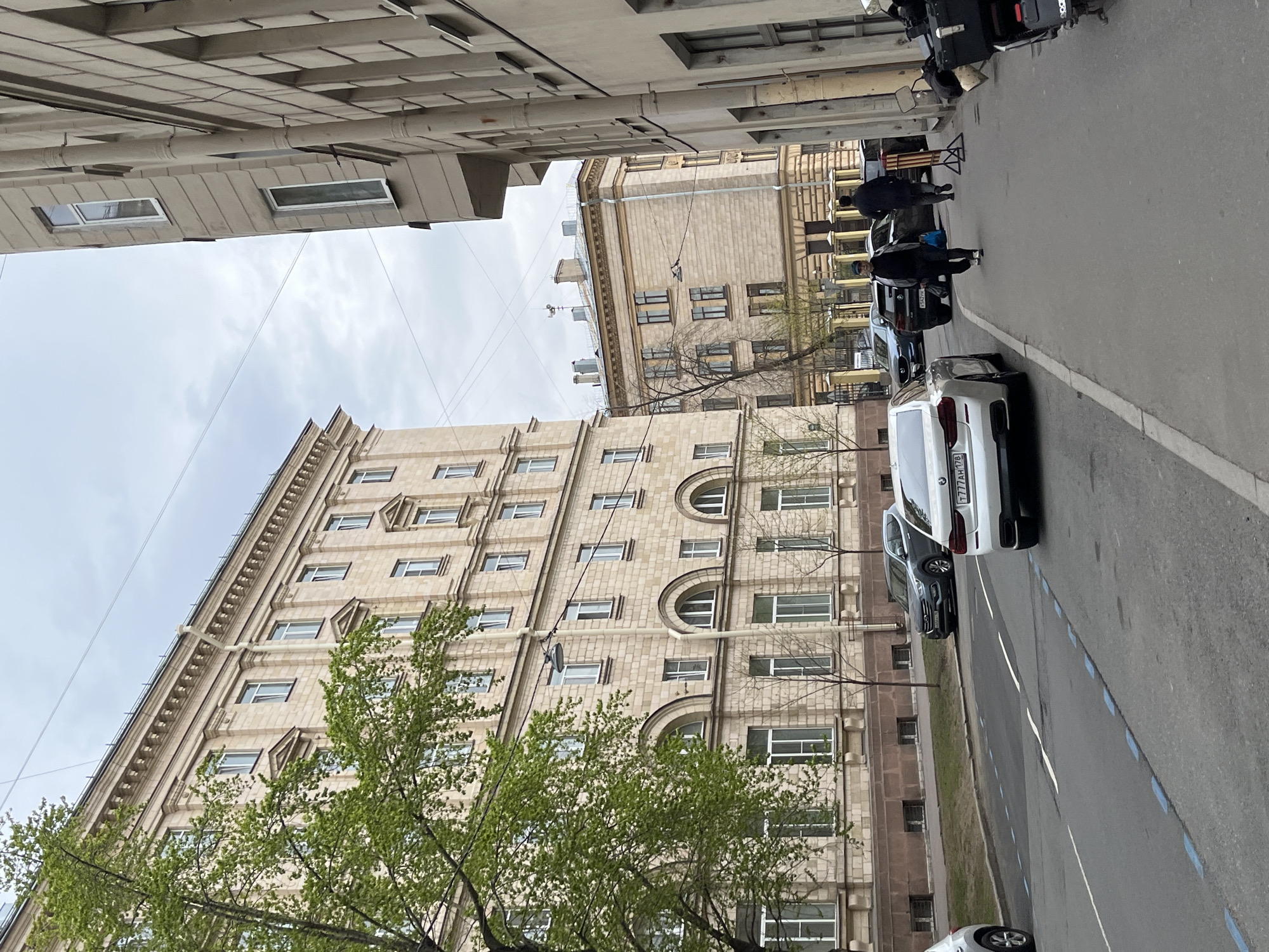 An eye-level outdoor shot depicts a street in Saint Petersburg, Russia, flanked by buildings and parked cars. The view is taken from the road level, angled slightly upwards to capture the architecture.
On the right, a line of parked cars extends into the distance. The first car in line is white and partially visible. Further ahead, people are walking on the sidewalk near the parked cars.
On the left side of the frame, a large, light-colored building with multiple windows dominates the scene. A green tree partially obscures the lower portion of the building.
The sky is overcast, and the overall lighting is soft and diffused.