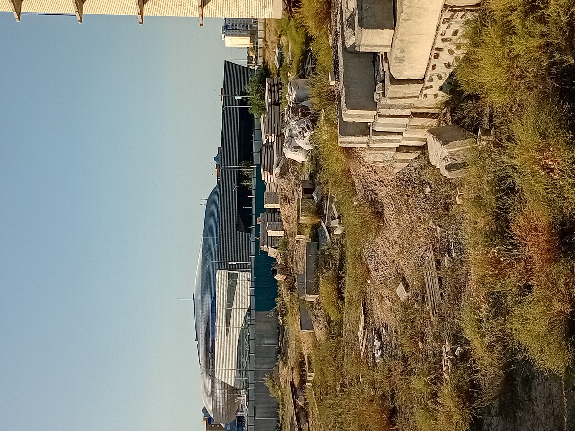 The image captures an outdoor scene in the city of Semey, Kazakhstan, dominated by a construction site in the foreground and a modern building in the background. The foreground is cluttered with building materials. Piles of concrete blocks are stacked, some forming a partially constructed wall. Several white sacks, possibly filled with cement or other construction materials, are present. Scattered debris, including loose stones and what appear to be discarded wooden planks, is spread across the uneven, overgrown ground. Dry, yellowing grass covers much of the foreground, indicating a dry climate or season. A concrete mixer is visible amongst the debris.
In the mid-ground, a metal fence separates the construction site from a larger area. Beyond the fence, a distinctive modern building stands out. It features a large, curved, metallic roof with a sleek, futuristic design. The building's facade appears to be a combination of glass and metallic panels with horizontal linear elements. The sky above is clear and blue, suggesting fair weather. To the far right of the image, a portion of an unfinished brick building is visible, with concrete supports protruding from its structure, further emphasizing the ongoing construction in the area. The overall impression is one of ongoing development and a stark contrast between the raw materials of construction and the polished appearance of the completed structure.