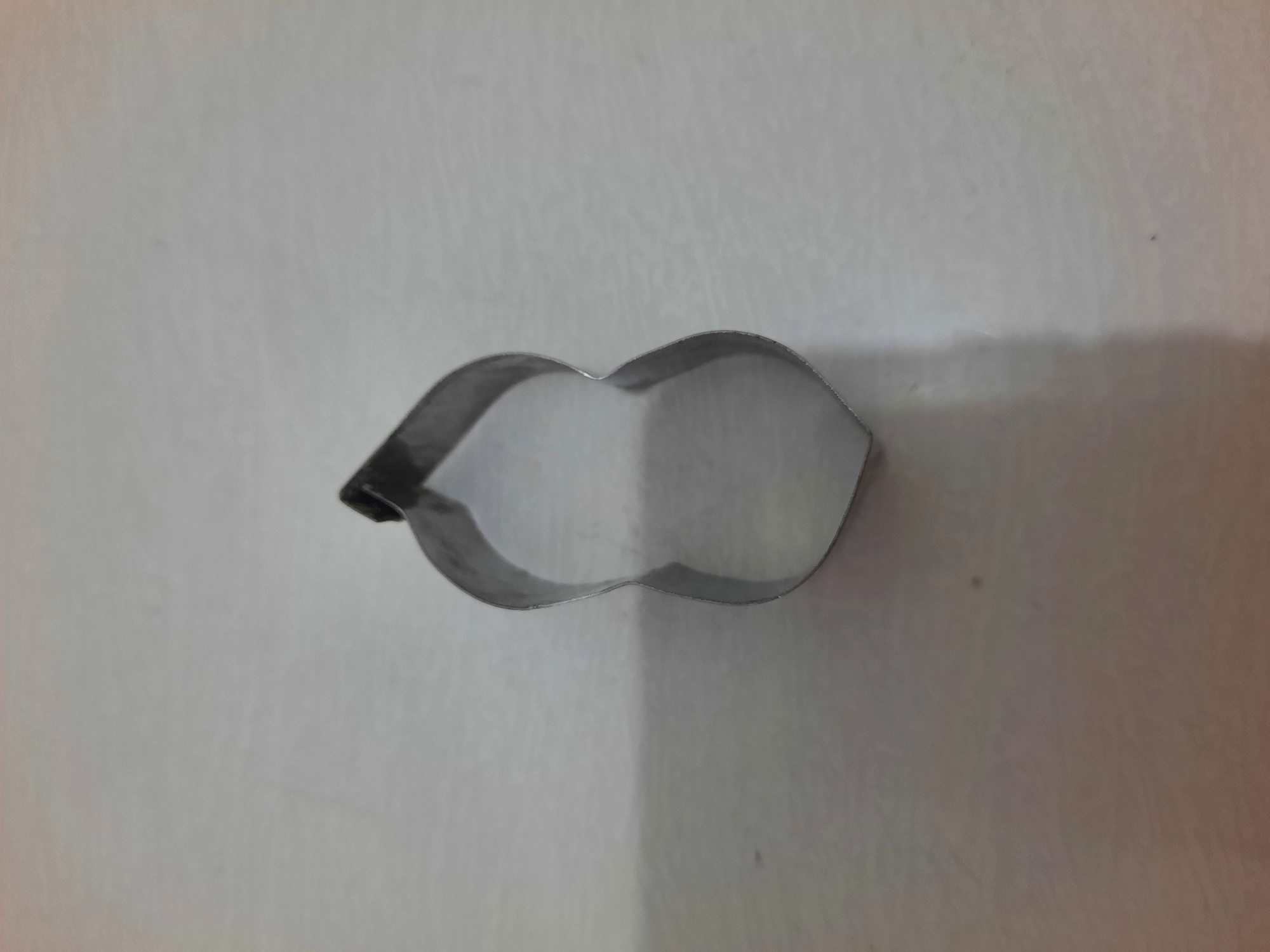 A close-up, slightly overhead shot shows a metallic cookie cutter lying on a light-colored, textured surface. The cookie cutter is shaped like a peanut, with two rounded lobes connected by a narrower middle section. The metal appears slightly worn and has a dull sheen. The background is a neutral, uniform color, suggesting an indoor setting, potentially a kitchen counter. There are no visible people or any other objects in the frame. The lighting creates a soft shadow on the surface behind the cookie cutter.