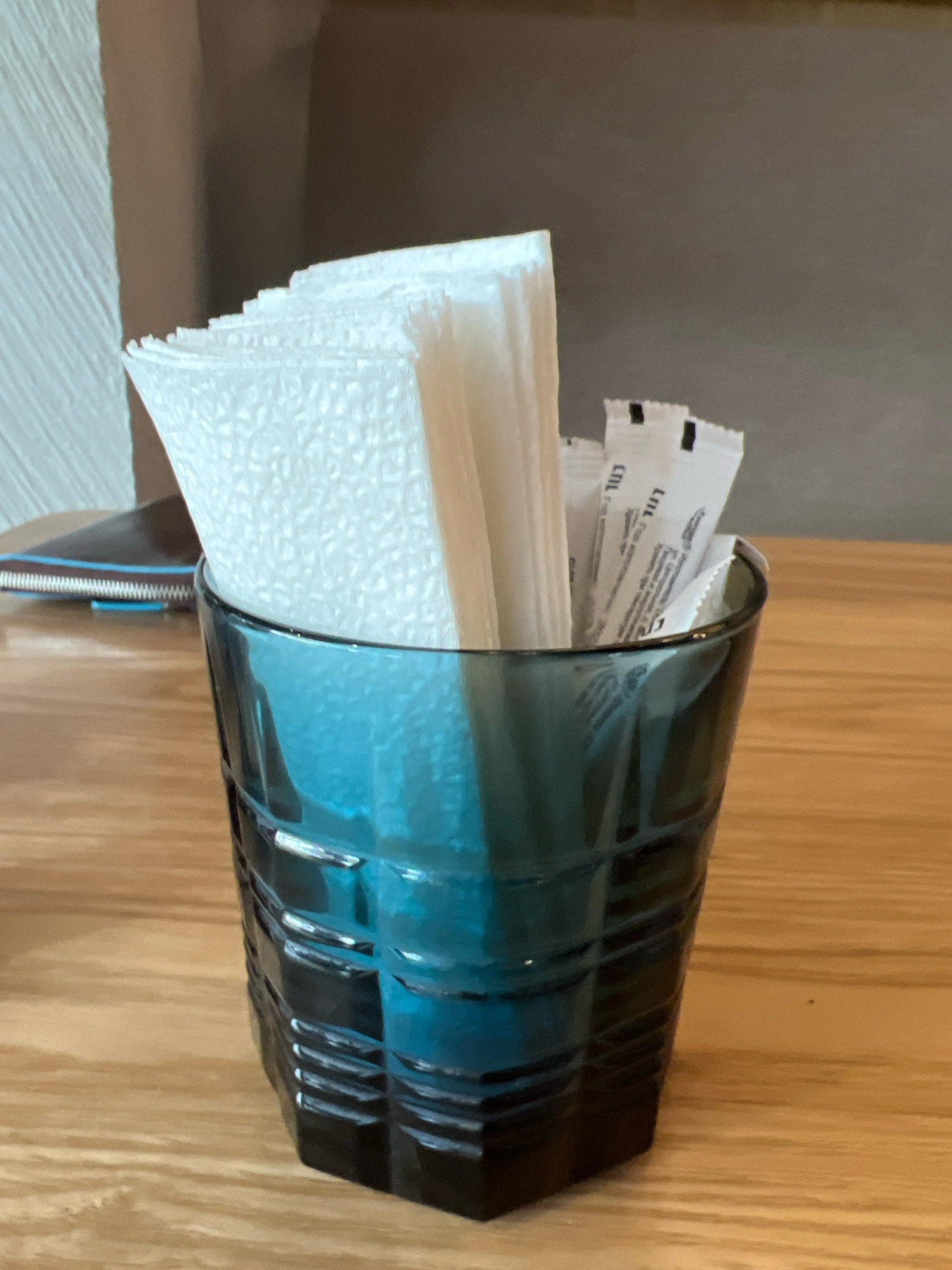 A close-up shot captures a blue glass tumbler filled with folded paper napkins and several packets of sweetener on a wooden table. The napkins have a textured pattern. The packets are white with black text and a black band at the top. The tumbler is faceted and appears to be made of dark blue glass. The background is a neutral brown, suggesting an indoor setting such as a cafe or restaurant. There is no indication of the specific location within the city of Krasnoyarsk, Russia, in this image.