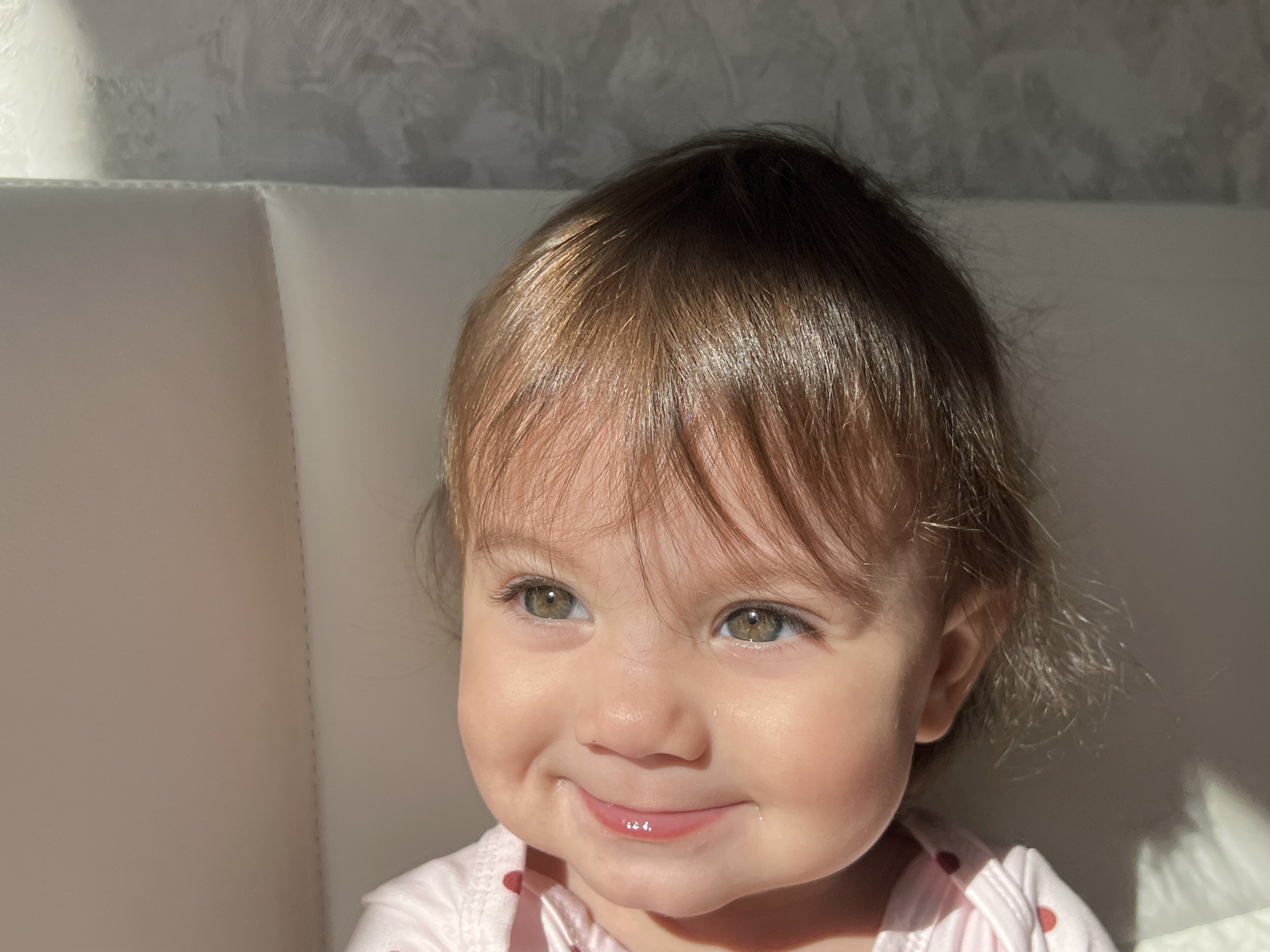 A baby with light brown hair and green eyes sits indoors against a white cushioned headboard. The baby is smiling, and wearing a light pink shirt. Sunlight illuminates the left side of the baby's face and casts shadows on the right. The environment suggests a room with soft, natural lighting. The image does not contain sufficient information to determine if the photo was taken in Elena, Bulgaria.