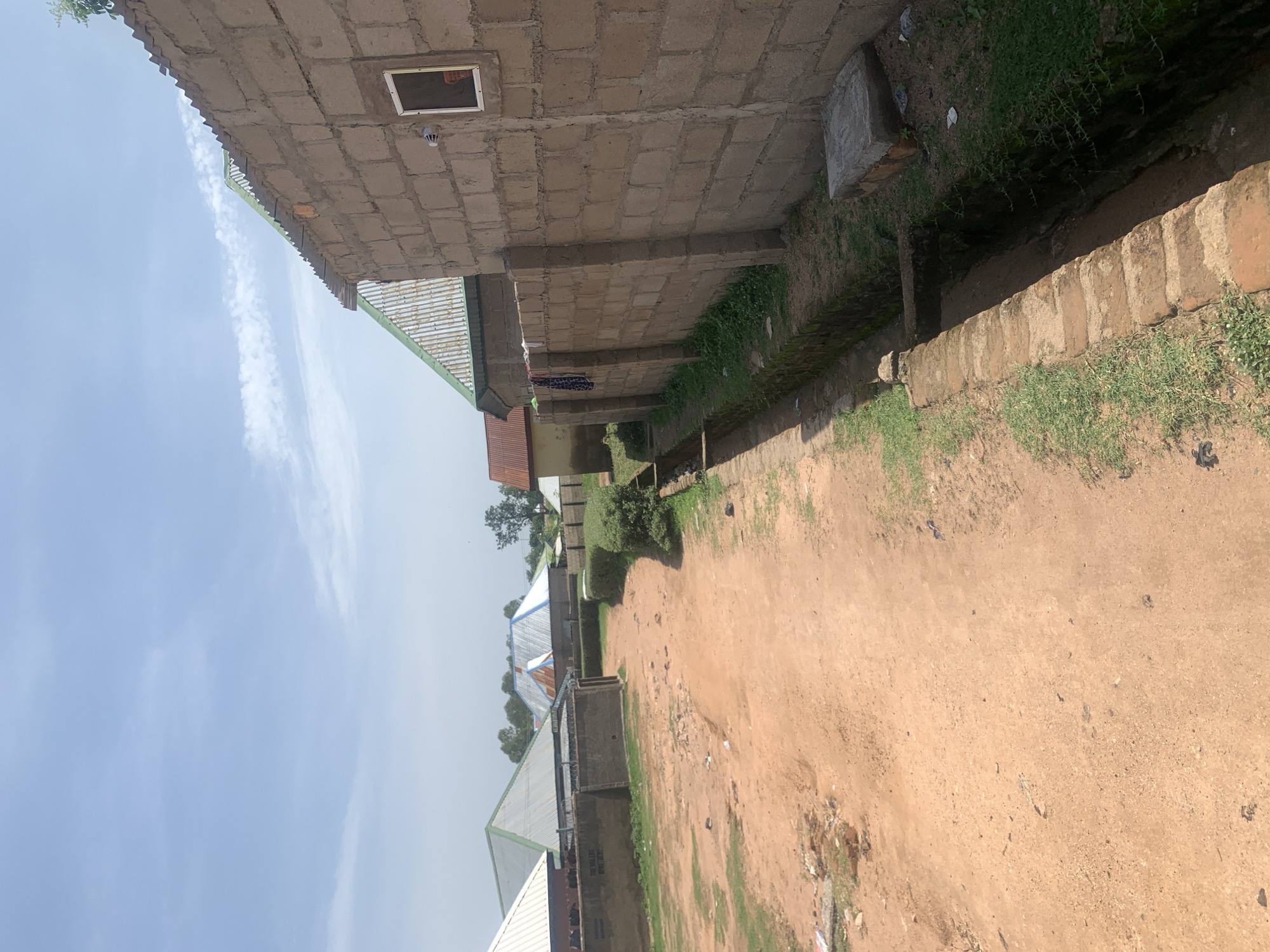 The image depicts a street scene in Jauro Yinu, Nigeria. A dusty, unpaved road runs diagonally from the bottom left towards the center of the image. On the right side of the road, a narrow, brick-lined drainage ditch is visible, with some overgrown grass. Adjacent to the ditch is a wall constructed from light-colored cinder blocks. A small, white outdoor light fixture is mounted on the cinder block wall, and a rectangular window with a white frame is set into the wall. Hanging from the wall are two pieces of dark fabric, possibly clothing. Further down the road, several buildings with corrugated metal roofs in varying colors (green, silver, and blue) are visible. These buildings appear to be residential or commercial structures. The sky above is a clear blue with a few wispy white clouds. The overall environment appears to be a somewhat developed but rural or semi-urban area.