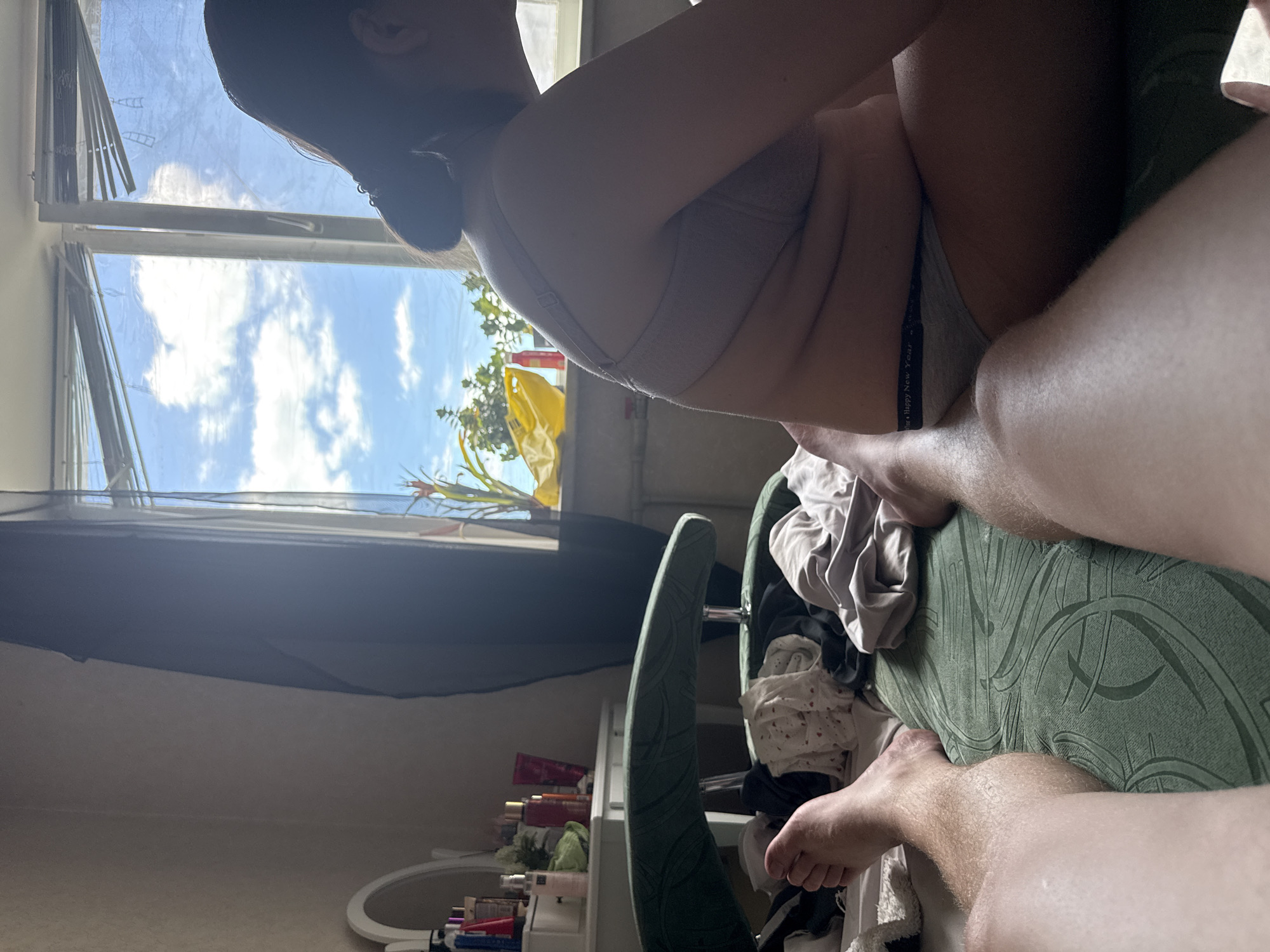 The image shows two people in what appears to be a bedroom. One person is partially visible from the side, wearing a bra and underwear. Their back is towards the viewer, and they are seated on a green chair. The other person's legs are visible in the foreground, also seated or lying down.
The scene is set near a window, through which a bright blue sky with white clouds is visible. Sunlight streams into the room, suggesting it is daytime. There are blinds on the window, and some greenery can be seen outside.
On a shelf below the window, several bottles of toiletries or cosmetics are arranged. A white framed mirror is also visible in the lower part of the image, along with some folded clothing or fabric. The overall impression is a casual, indoor setting, possibly a personal space.
The visible text on the underwear reads "Happy New Year". The mood is relaxed and intimate, suggested by the close proximity of the two individuals and their attire.