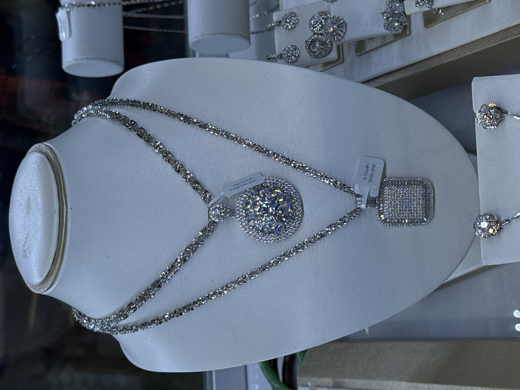 The image shows a close-up of a jewelry display, likely in Tay Nha Trang Ward, Vietnam, given the currency denomination (VND) on the price tag of 9,200,000 VND on the left and 1,680,000 VND on the right of the chain. The focus is on a silver-colored necklace with a round pendant. The necklace and pendant appear to be encrusted with small, clear stones (likely diamonds or cubic zirconia). The necklace is draped over a white, curved display stand. In the background, other jewelry pieces are visible, including earrings and possibly rings, also set with clear stones. The display suggests a retail setting, such as a jewelry store. The lighting is bright, highlighting the sparkle of the stones.