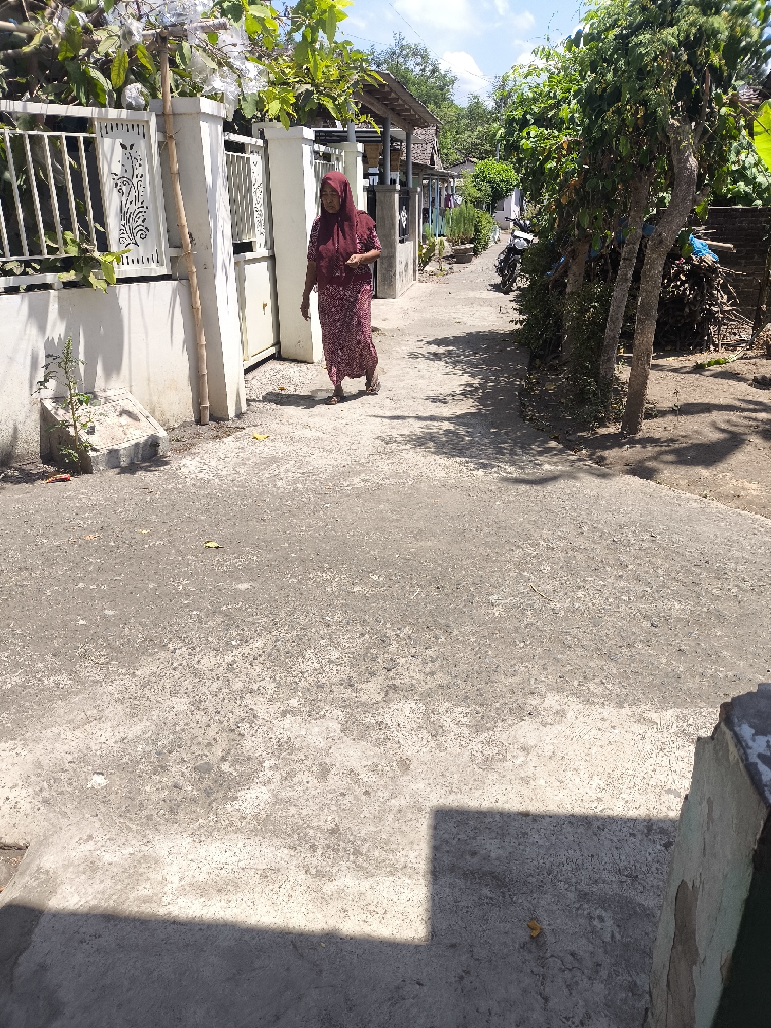 An elderly woman wearing a maroon hijab and a matching patterned dress walks on a paved path in a village in Grati, Indonesia. She is walking away from the camera, towards a white gate and fence on the left side of the image. To the right of the path are trees and a pile of wood. The scene is illuminated by bright sunlight, casting shadows on the ground. The path is made of concrete and appears worn. The buildings in the background are simple, with corrugated iron roofs.