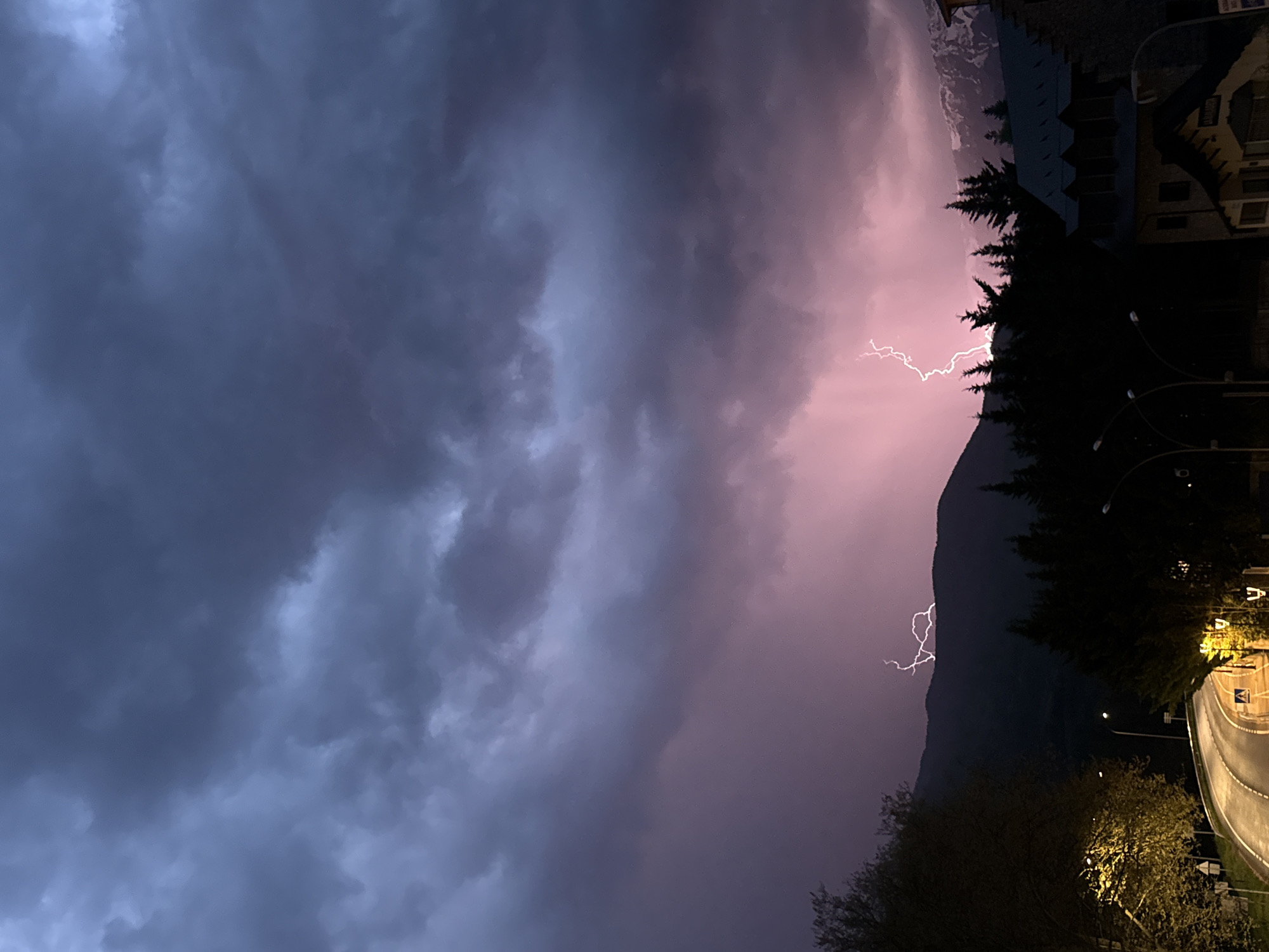 A dramatic nighttime photograph captures a powerful lightning storm over a mountainous landscape in Benasque, Spain. The upper portion of the image is dominated by heavy, dark grey and purplish storm clouds, indicating severe weather. A brilliant, jagged white lightning bolt dramatically forks across the sky and a mountain peak, illuminating the surrounding clouds with a striking pink and purple glow.
Below the tumultuous sky, the dark silhouette of a rugged mountain range is visible, partially obscured by clouds and dense evergreen trees. In the lower right of the frame, elements of a town or village are discernible. A curving road, illuminated by warm yellow streetlights, creates a bright path, and its light casts a glow on nearby trees and foliage. Buildings with dark roofs and some visible windows are nestled against the mountain on the far right. The overall atmosphere is one of intense natural power, with the lightning providing a stark contrast against the dark, stormy sky and the silhouettes of the landscape and urban structures. The scene clearly depicts a nocturnal event, highlighted by the darkness and artificial illumination. No discernible text is visible in the image.