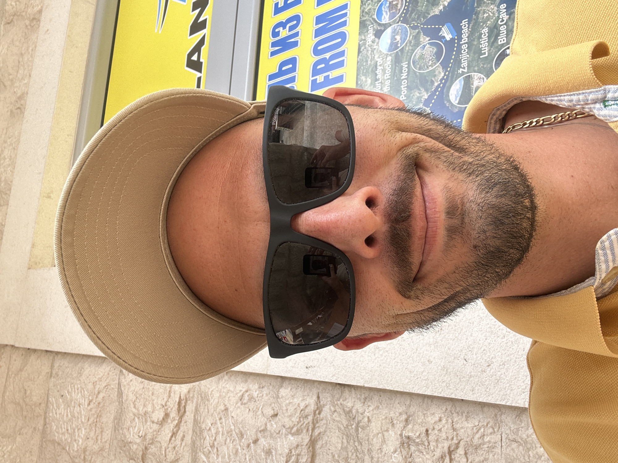 A man is depicted in what appears to be a selfie or close-up shot, angled vertically with his head on the left. He is wearing a beige baseball cap, black rectangular sunglasses, and a yellow polo shirt with a blue and white striped pattern visible on the collar. A gold chain is visible around his neck. He has a short, dark beard and is looking towards the camera with a subtle smile.
The background features a large yellow sign with blue text, partially visible, which reads "LAN", "ЛЬ ИЗ Е", and "FROM". Below this, there's a map or poster with several location names printed on it, suggesting tourist attractions or tour destinations. These include "Jur Lady of the Rocks", "Porto Novi", "Risan", "Žanjice beach", "Luštica", and "Blue Cave". The sunglasses reflect what looks like another person holding a phone, indicating the picture might be a selfie. The brand "Polaroid" is visible on the sunglasses frame. The ground or a nearby wall is light-colored and textured. The bright lighting suggests it is daytime. The presence of these specific locations on the map aligns with the provided context of Budva, Montenegro, as these are popular sites in the Bay of Kotor and its vicinity. The scene suggests a tourist activity, possibly related to boat tours or excursions.