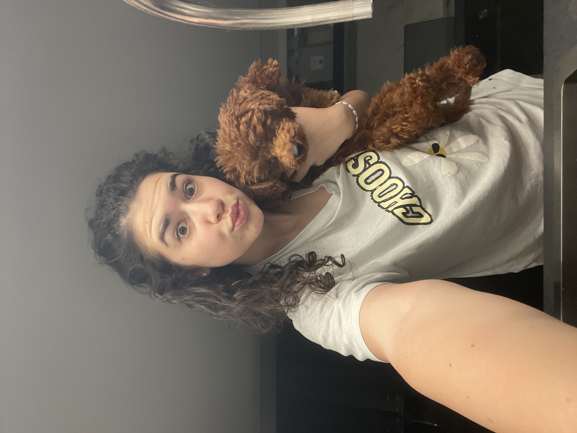 A woman is holding a brown poodle close to her face, as if taking a selfie. The woman has dark, curly hair, fair skin, and is wearing a white t-shirt with the word "SOOKS" printed on it. She is making a duck-face expression. The dog is nestled against her shoulder and appears to be looking at the camera. The background appears to be an indoor scene, possibly a kitchen or bathroom, based on the presence of a metallic faucet-like fixture in the upper left corner. The image is a close-up shot. There is no immediate indication of the location being Sofia, Bulgaria.