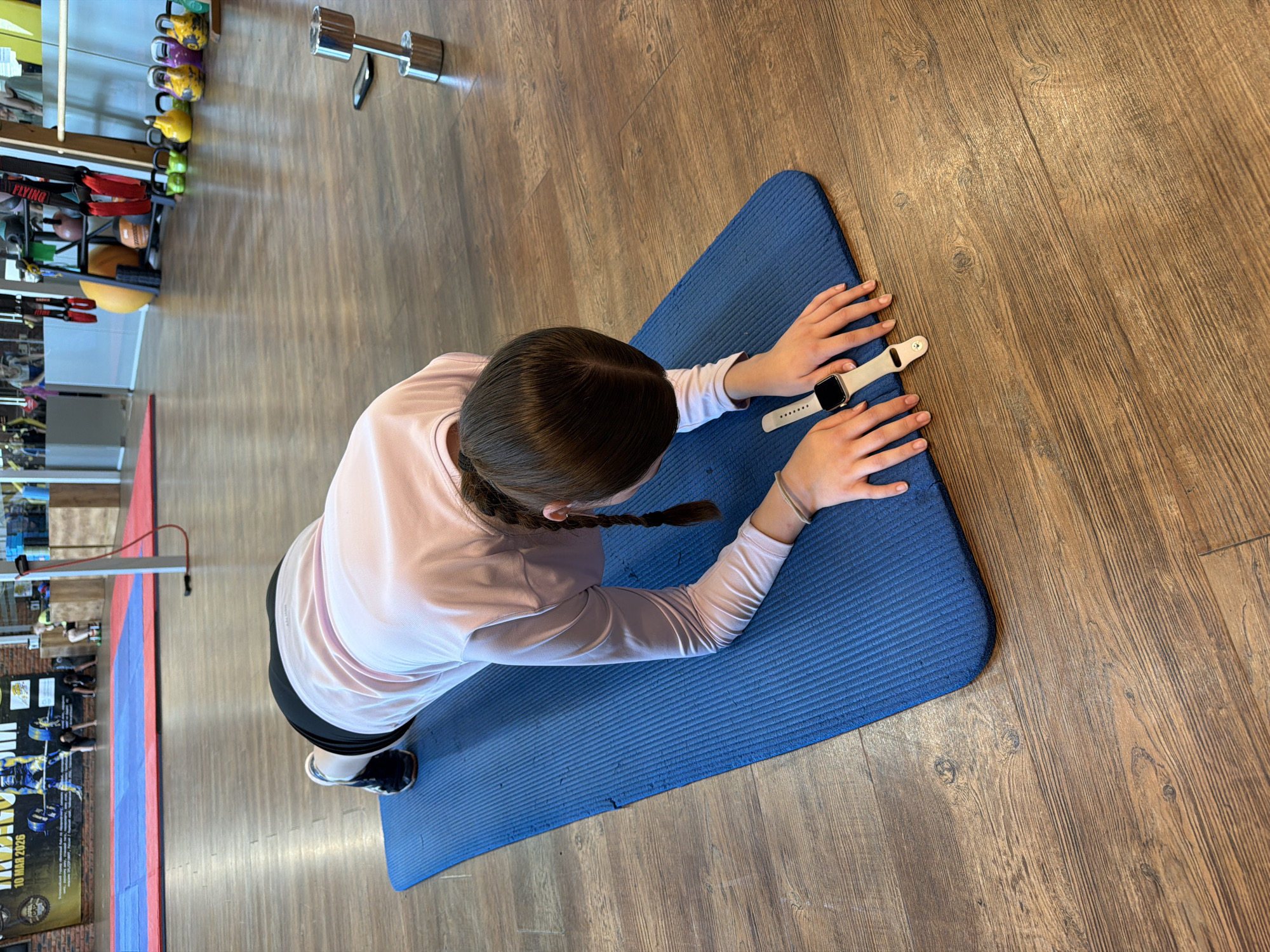 A young person with braided hair is performing a plank exercise on a blue yoga mat on a wooden floor in a gym. They are wearing a light pink long-sleeved shirt and black shorts. A smartwatch is visible on their left wrist. In the background, weightlifting equipment, including dumbbells and kettlebells, is present. The scene is indoors, likely in a sports facility located in Chaikovsky, Russia, as suggested by the banner in the background that mentions "CHAIKOVSKY" and "10 MAR 2026".