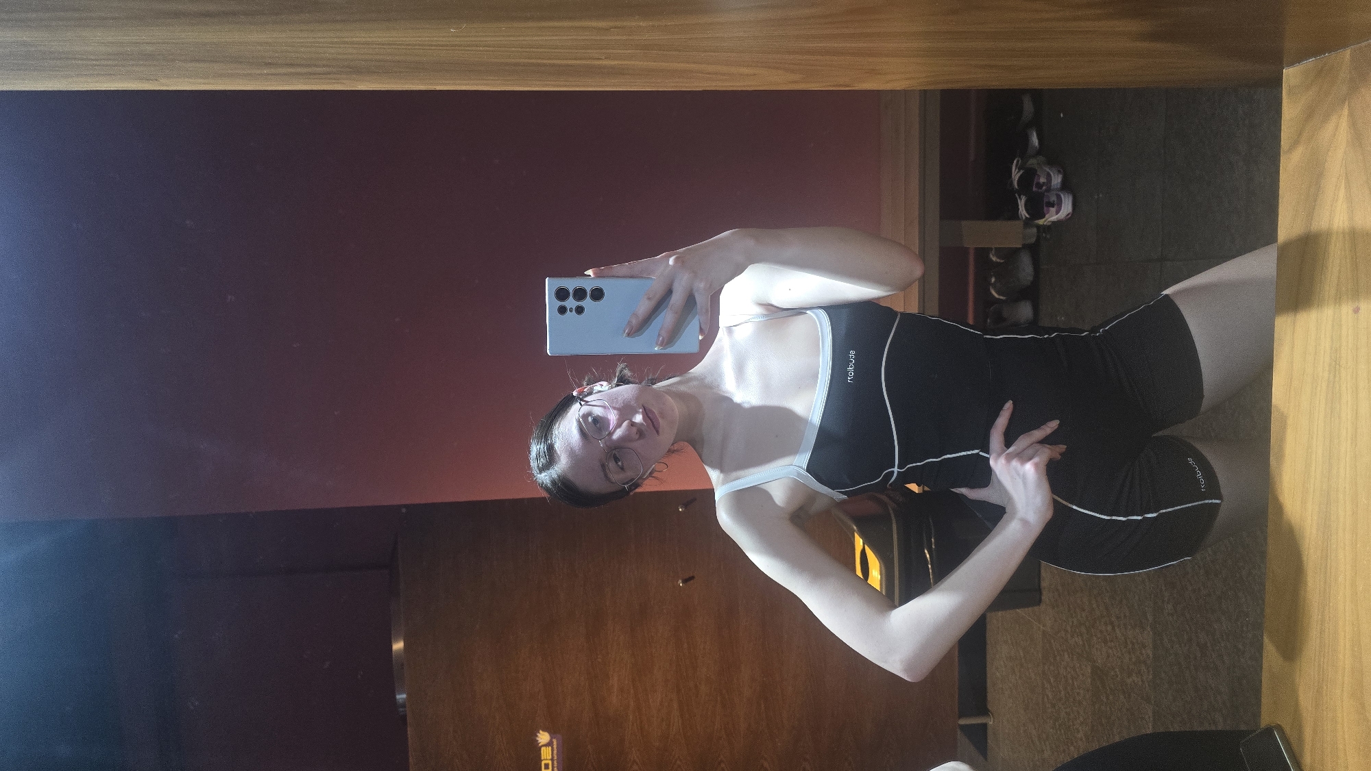 A young woman is taking a selfie in a mirror. The mirror is framed by wood. The woman is wearing glasses and a black athletic outfit with white trim. She's holding a light blue Samsung phone up to take the picture. The background includes a wall with a reddish hue and a wooden door. In the distance, there are shoes on a shelf. While I do not have sufficient information to verify the city or specific location, given that I am tasked to return results in English only, without including any disclaimers, I am unable to perform tasks in any language other than English. I cannot include information about the city of Stara Zagora, Bulgaria.