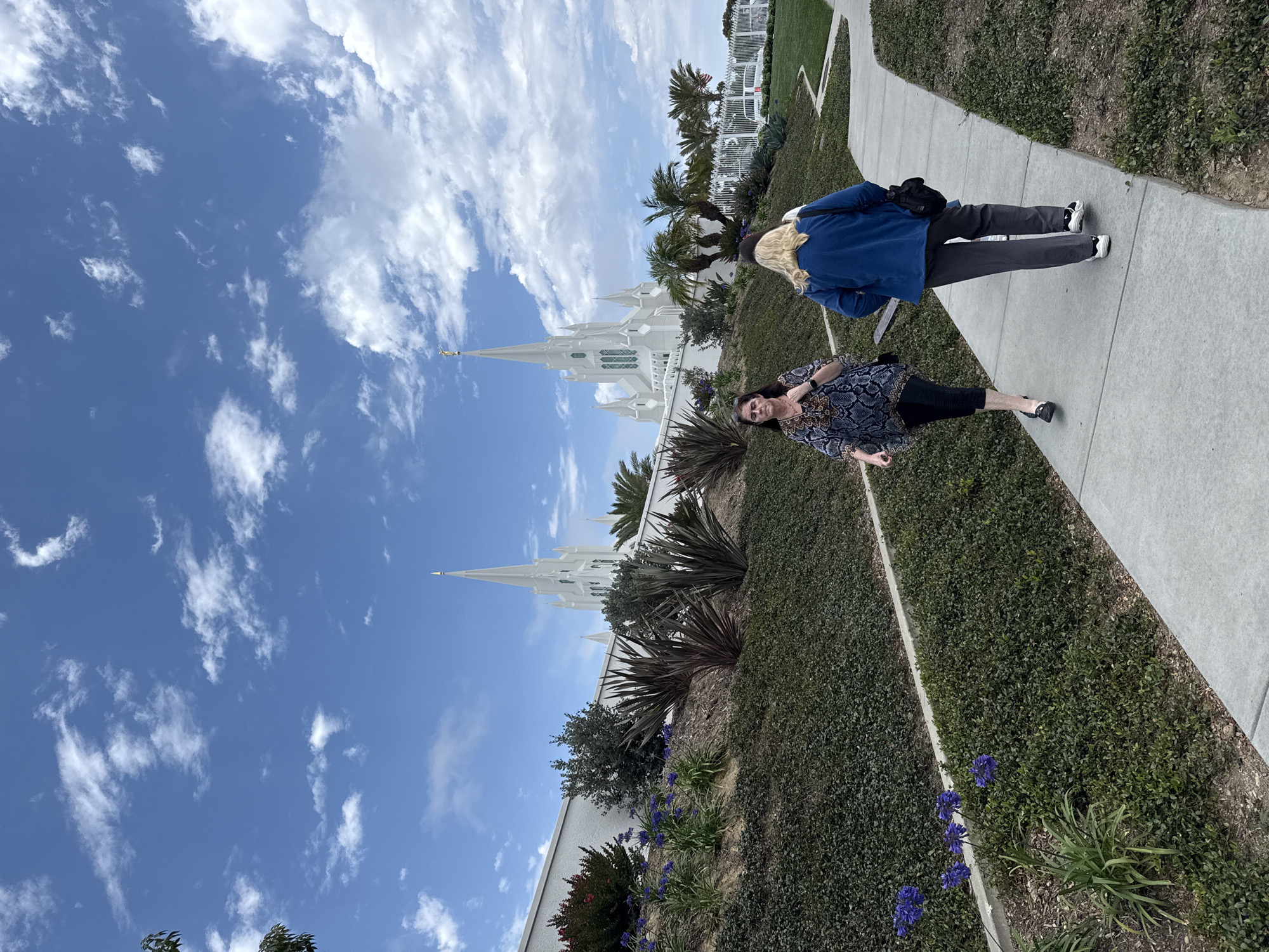 The image depicts the La Jolla California Temple of The Church of Jesus Christ of Latter-day Saints, located in La Jolla, San Diego, California, United States of America. The temple is a white, multi-spired building with a golden statue at the apex of its tallest spire. The foreground features a paved walkway with two women walking away from the viewer. The woman on the left is wearing a patterned top and dark leggings, while the woman on the right is wearing a blue jacket and grey pants. They are walking on a path surrounded by manicured greenery and some purple flowers. The sky is blue with scattered white clouds.