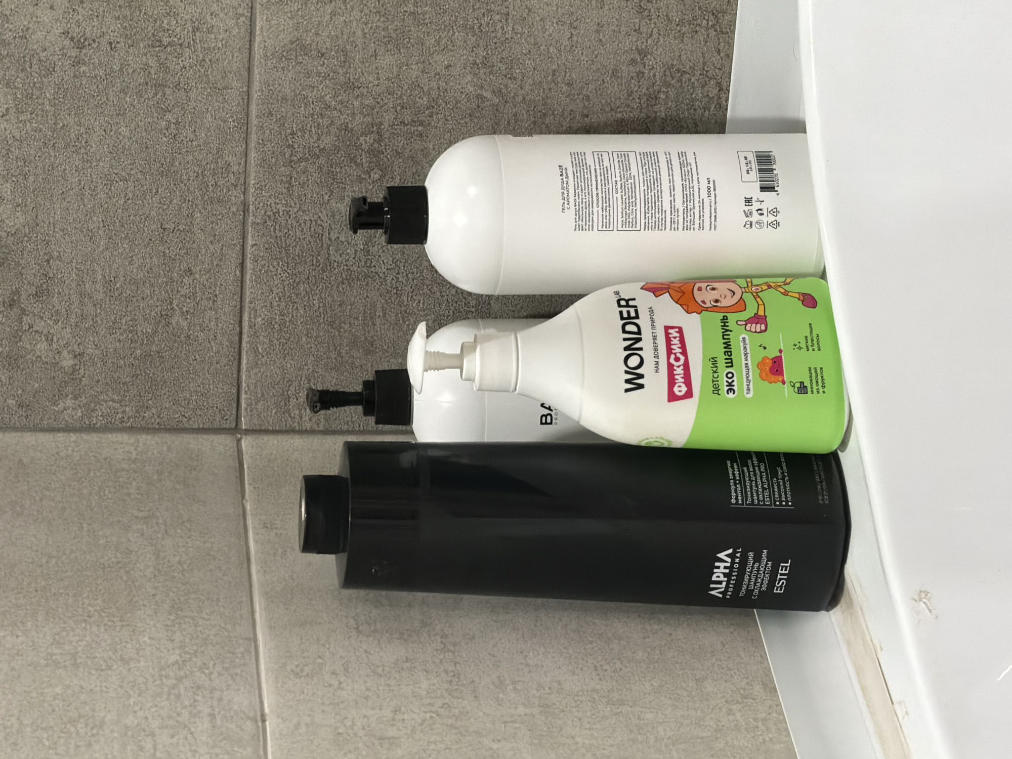 The image depicts a static close-up of four plastic bottles positioned on a white corner shelf against a tiled wall in what is presumed to be a bathroom in Chaikovsky, Russia. The wall consists of two distinct gray tile types: a lighter, smoother textured tile on the left and a darker, granular textured tile on the right, separated by a vertical grout line. A horizontal grout line bisects both tile sections.
From left to right, the bottles are:
1. A tall, black, rectangular bottle with a black cap, labeled "ALPHA PROFESSIONAL ESTEL," identified as a "ТОНИЗИРУЮЩИЙ ШАМПУНЬ С ОХЛАЖДАЮЩИМ ЭФФЕКТОМ" (Tonic Shampoo with Cooling Effect), formulated with menthol and caffeine.
2. A partially obscured white cylindrical bottle with a black pump dispenser, bearing the partial brand name "BA".
3. A prominent green and white bottle with a white pump dispenser, labeled "WONDER" and featuring "ФИКСИКИ" (Fiksiki) branding. This bottle contains "ДЕТСКИЙ ЭКО ШАМПУНЬ" (Children's Eco Shampoo) and depicts a cartoon character, promoting soft, shiny hair and natural ingredients.
4. A large white cylindrical bottle with a black pump dispenser, identified as "ГЕЛЬ ДЛЯ ДУША BAZE С АРОМАТОМ ДЫНИ" (Shower Gel BAZE with Melon Scent). Its label details product information, ingredients (including Aqua, Sodium Laureth Sulfate, Cocamidopropyl Betaine), application instructions, manufacturer details, a 1000 ml volume, and a visible date "26.12.20."
The white shelf exhibits minor surface discoloration and small chips along its front edge. No individuals or active interactions are present in the scene.