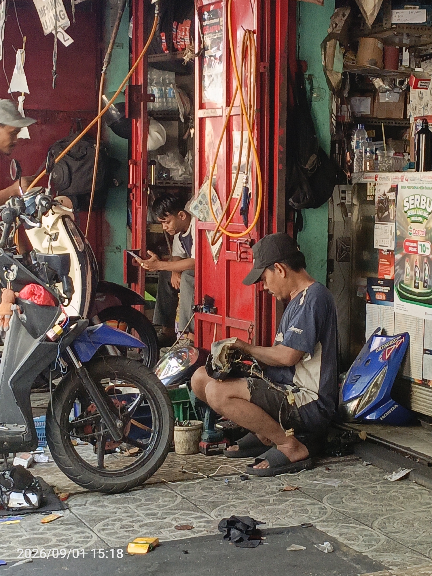 The image depicts an outdoor scene in East Jakarta, Indonesia, likely a motorcycle repair shop or a street-side vendor. In the foreground, a man wearing a black baseball cap and a blue t-shirt is squatting on the tiled ground, working on disassembled motorcycle parts. He is wearing dark shorts and black sandals. To his left, a partially visible motorcycle with cream-colored bodywork and a blue front fender is parked. In the background, another man in a blue and white shirt is standing and looking at a mobile phone. Behind him, red doors and shelves are visible, stocked with various items including bottles and what appear to be spare parts or products. The environment appears cluttered, with tools, wires, and debris scattered on the ground. A Honda logo is visible on a blue motorcycle fairing to the right of the man working. The overall impression is one of a busy, functional workshop.