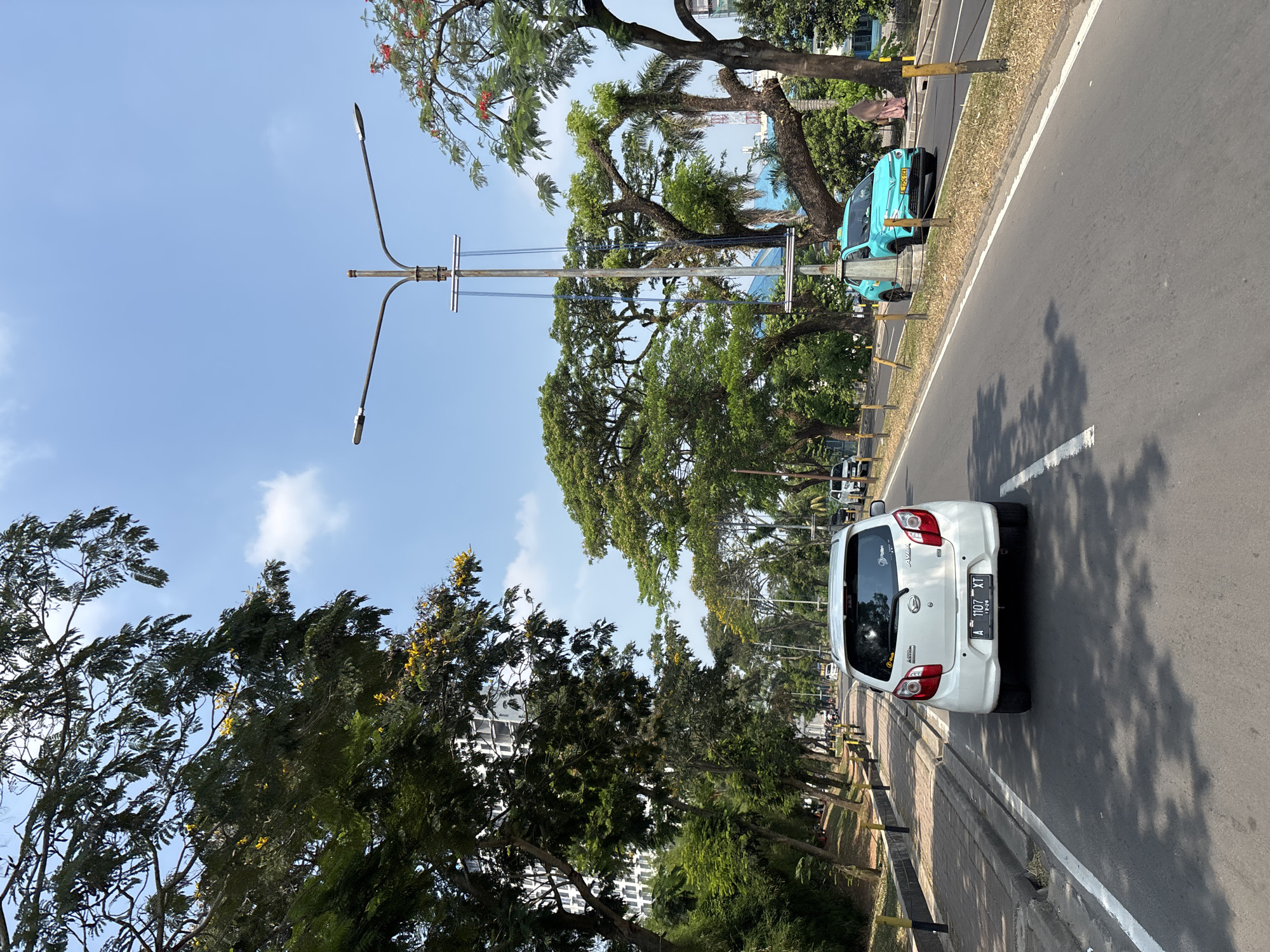 The image shows a street scene in Bencongan Indah, Indonesia, on a bright, sunny day with a clear blue sky and scattered clouds. The viewpoint is from a vehicle traveling on the right side of the road. In the foreground, a white car is visible from the rear, with its license plate reading "A 1107 XT". Adjacent to the road on the left is a sidewalk with patterned paving stones and yellow and black bollards. A line of trees with lush green foliage, some bearing small yellow flowers, borders the sidewalk. Beyond the trees, a tall, multi-story building with a grid-like facade is partially visible.
The road itself is asphalt, with a white lane marker. Further ahead on the right side of the road, a turquoise car is in motion. This car has a license plate starting with "B". In the background, more trees with dense green canopies and some with red flowers are prominent. A street light pole with two lamps stands near the turquoise car. A few other vehicles are sparsely distributed on the road. In the far background, several buildings and structures, including what appears to be a tall tower with scaffolding, are visible. The overall environment suggests a well-maintained urban or suburban area with significant greenery.