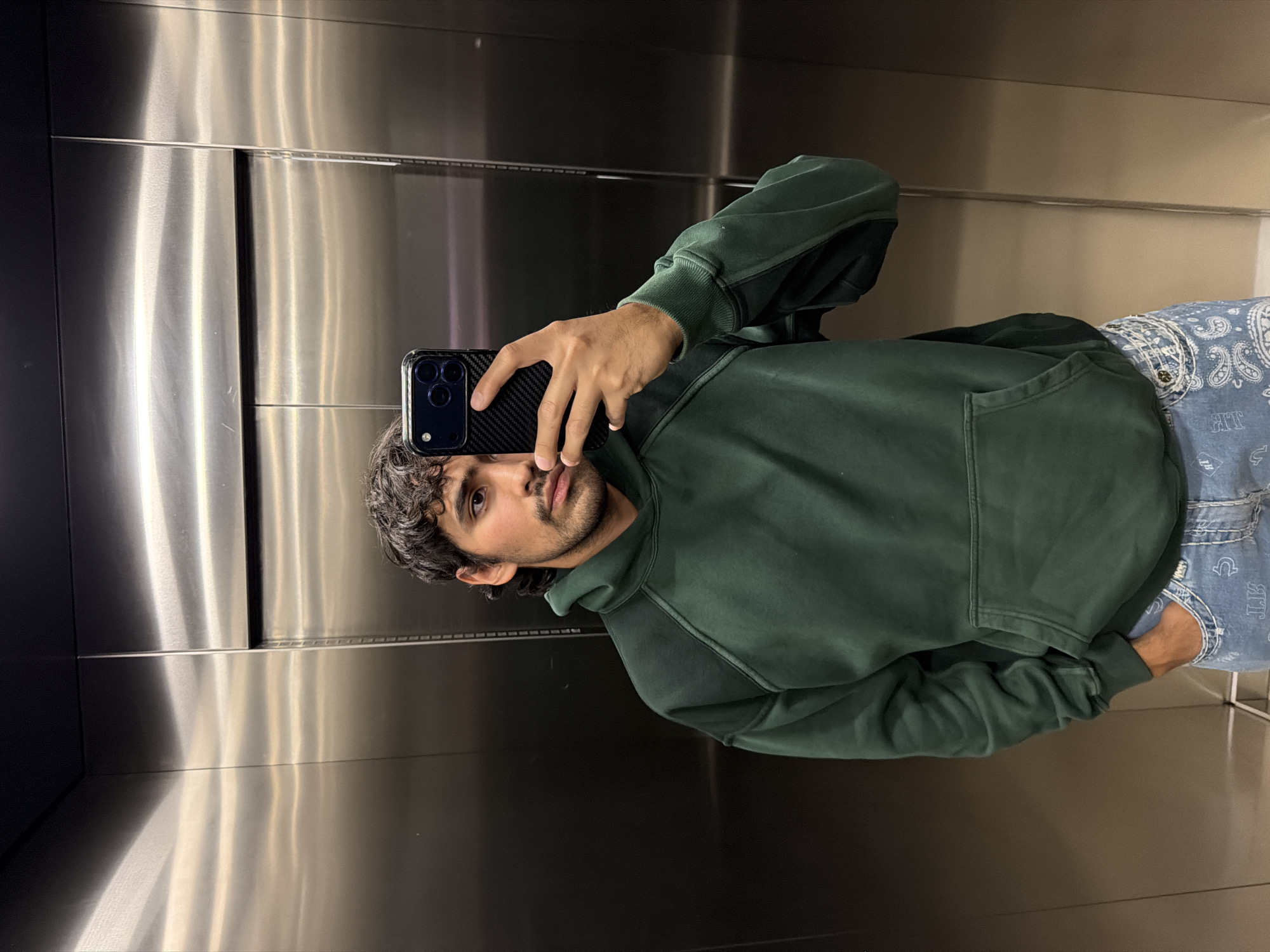 A person is taking a selfie in what appears to be an elevator with a metallic interior. The person has curly black hair and a beard. They are wearing a dark green hoodie and denim jeans. The phone being used to take the picture has a black case with a three-lens camera system. The setting is not definitively located in San Andrés Cholula, Mexico, and the elevator’s design does not indicate its location.