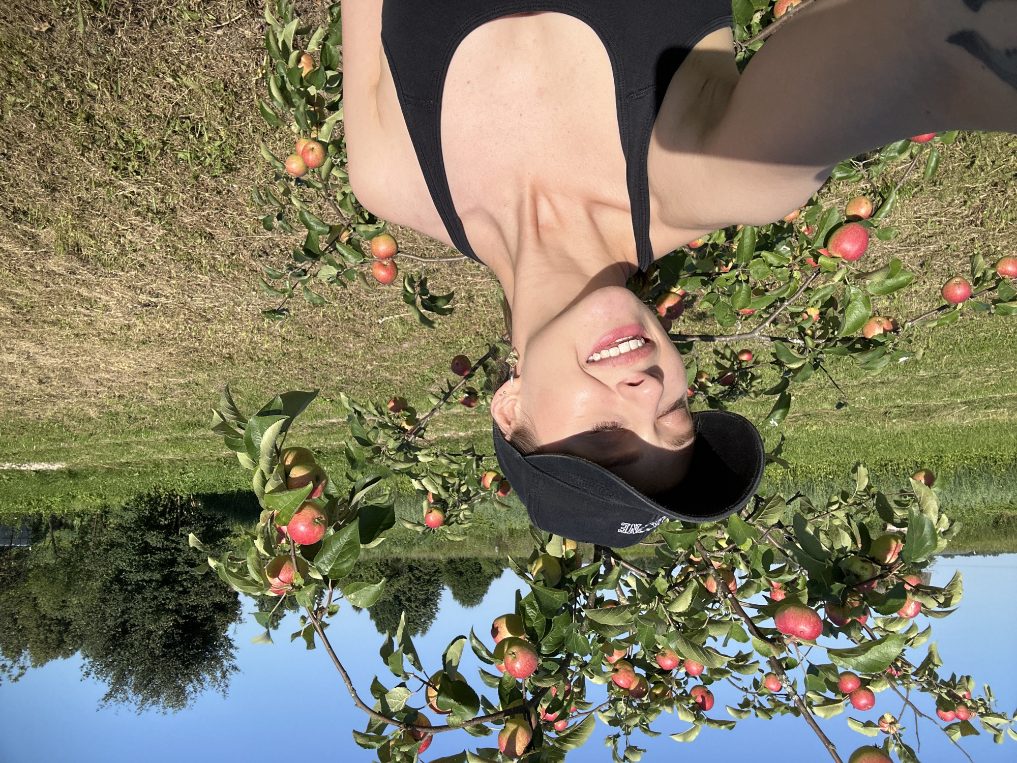 A woman in a black baseball cap and tank top smiles with her eyes closed while standing in front of an apple tree laden with red apples. The setting is outdoors, with a blue sky and green grass visible. The background includes trees and a field. The image appears to be a selfie taken on a sunny day. The location is Turovo, Russia.