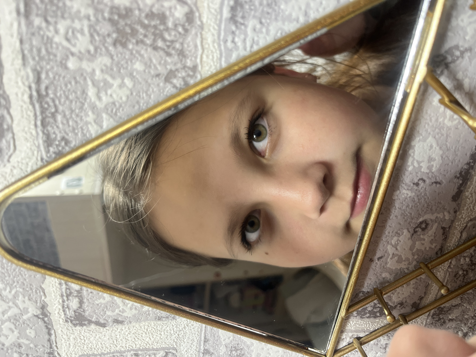 A girl's face is reflected in a triangular, gold-framed mirror. The background shows a patterned wall. The location is not discernible.