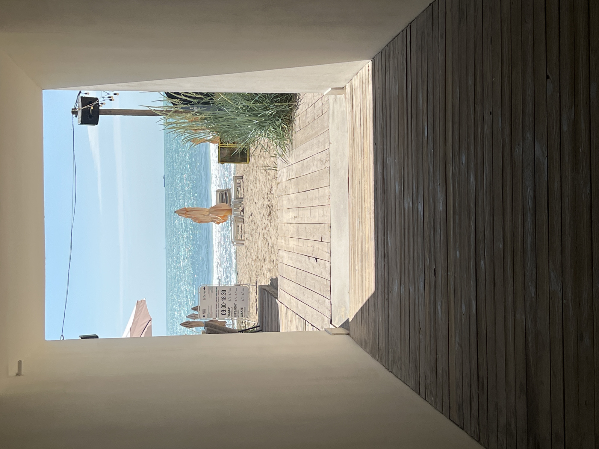 The image captures a serene beach scene, viewed through a concrete archway or opening that frames the composition. The perspective is from an indoor or covered area, looking out towards the sea.
In the foreground, a light-colored wooden walkway leads from the viewer's position down a gentle slope towards the sandy beach. To the right of the walkway, within the frame, is a dark wooden slatted wall, suggesting part of a building or structure. To the left, a light concrete ledge is visible, with a small dark object (possibly a phone) resting on it.
The middle ground features a wide expanse of light brown sand. Several light beige or tan folded beach umbrellas are scattered across the sand, alongside some stacked lounge chairs, indicating a beach resort or serviced area. Further back, a patch of tall green reeds or grasses grows on the sand, behind which a yellow metal structure is partially visible. Above this area, a dark speaker is mounted on a pole, from which string lights hang.
The background is dominated by the calm, sparkling blue sea under a clear, bright blue sky. Gentle waves break near the shore. There are no people visible in the scene.
A white rectangular sign with black text is prominent on the beach. It reads: "РАБОТНО ВРЕМЕ / WORKING HOURS" followed by "09:00 - 18:30". Below this, a list of items appears in Cyrillic script with associated prices, including "ЧАДЪР" (umbrella), "ШЕЗЛОНГ" (sunbed/lounge chair), and "ШАТРA" (tent/canopy), indicating services available at the beach.
The scene is brightly lit by natural sunlight, suggesting a clear day, likely morning or early afternoon, with shadows cast by the structures. The overall atmosphere is peaceful and inviting, characteristic of a pleasant day at the beach in Varna, Bulgaria.