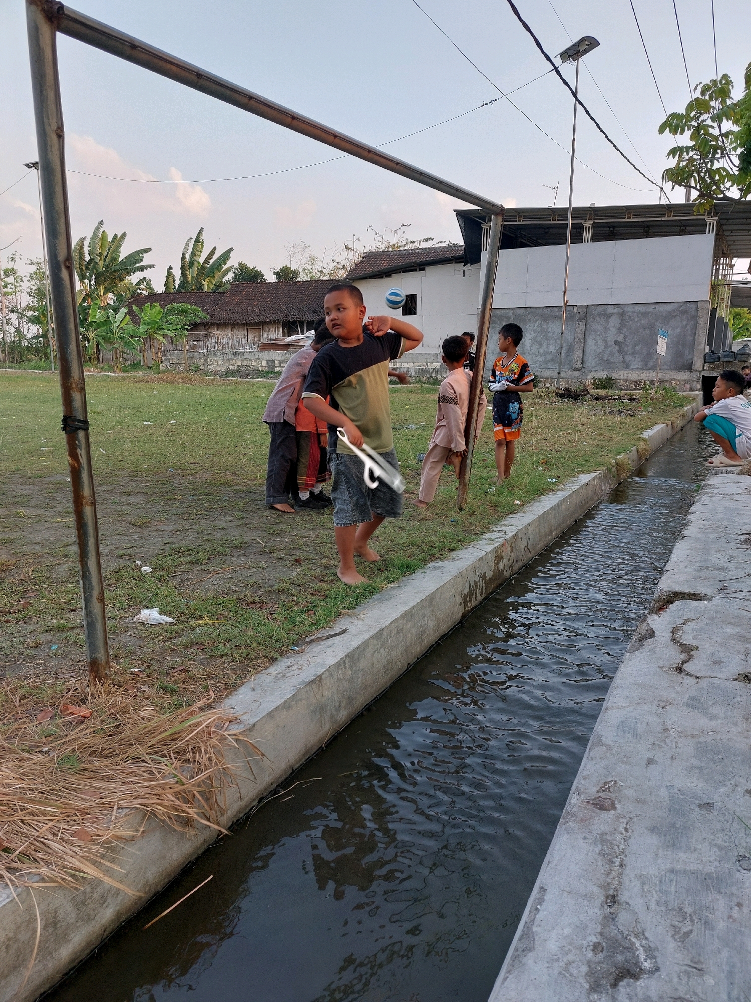 In Sarangan, Indonesia, a group of children is gathered in an outdoor area adjacent to a concrete irrigation ditch. The ditch is filled with dark, rippling water. On the grassy field beside the ditch, several children are present. One boy in the foreground, wearing a dark t-shirt with a green and yellow diagonal stripe and denim shorts, is holding a metal object that appears to be a wrench or similar tool. He is looking towards the right of the frame. Behind him, a few other children are standing or leaning against a wooden pole. Further in the background, a building with a tiled roof and a white facade is visible, along with several banana trees and other vegetation. A tall pole with a solar-powered light is also prominent in the scene, with power lines strung overhead. The sky is partly cloudy.