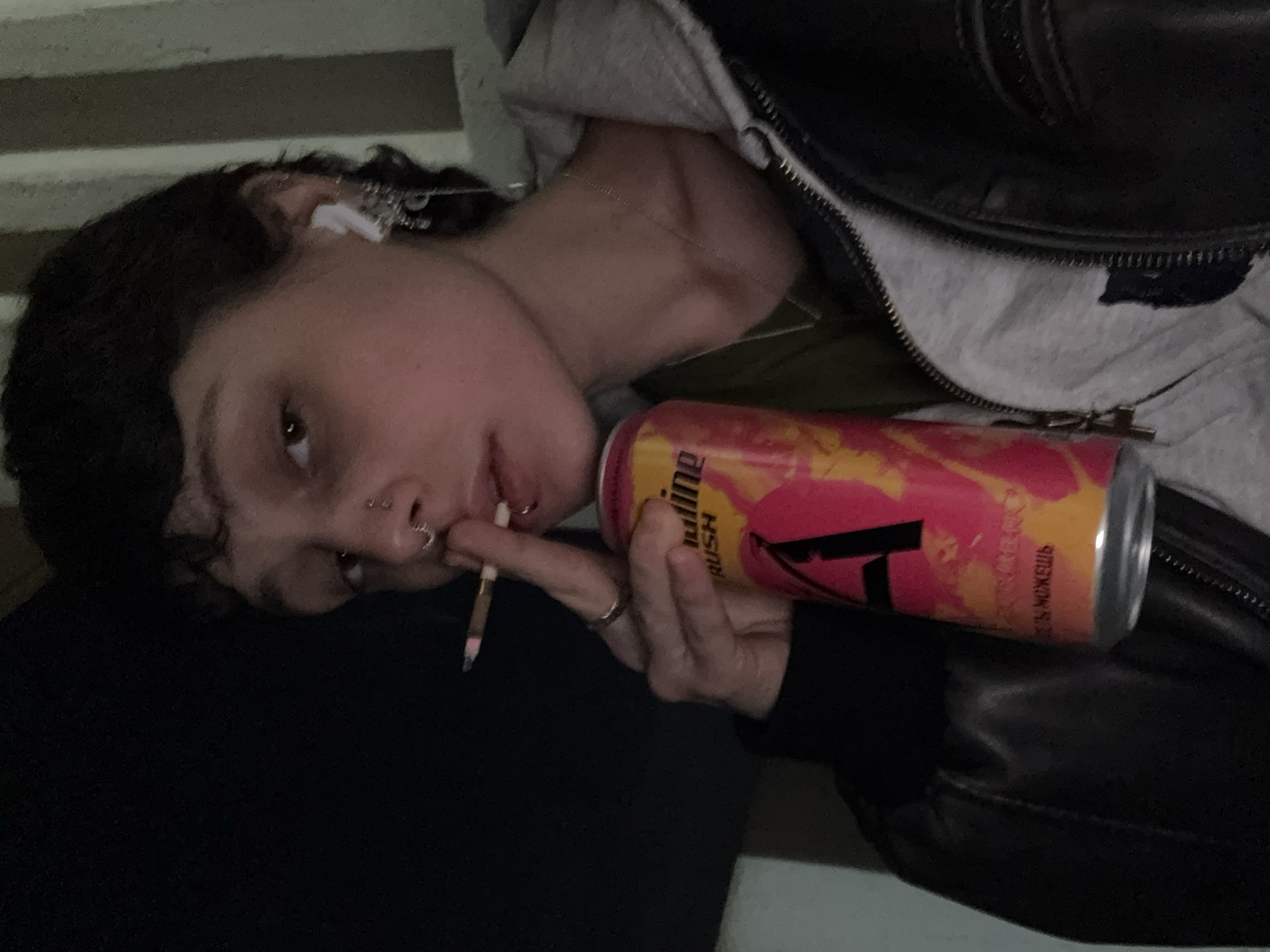 A person with a cigarette in their mouth leans against a dark surface, holding a pink and yellow energy drink can. The can has the Cyrillic alphabet written on it. The person is wearing a gray hoodie and a black leather jacket. The location is likely Moscow, Russia, given the Russian text on the can. The person has multiple piercings in their nose and is wearing earbuds. The background shows a glimpse of a textured wall.