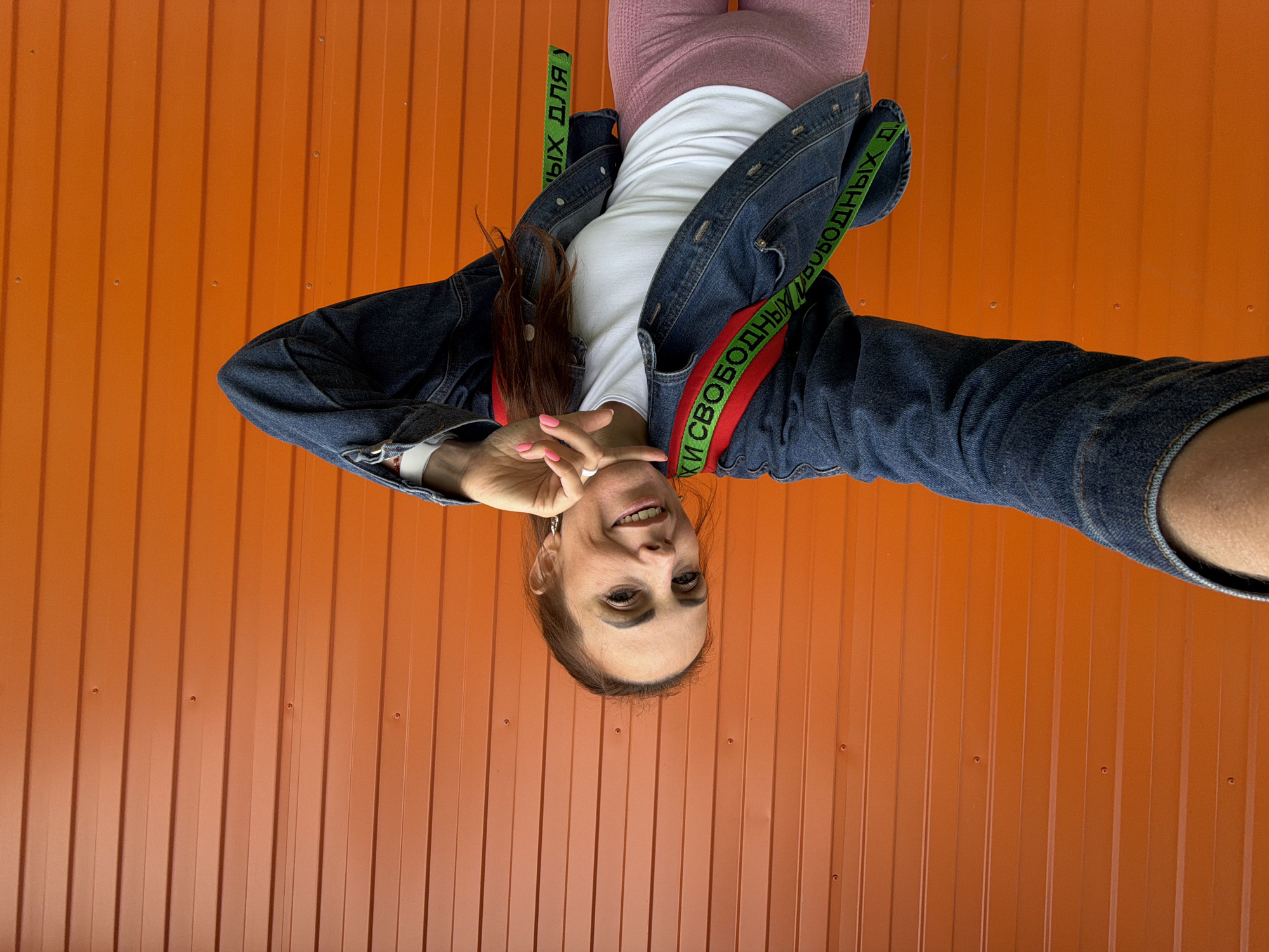 A woman with long brown hair, wearing a denim jacket over a white t-shirt and pink leggings, stands in front of an orange corrugated metal wall. A red backpack with a green strap, featuring black text, is visible on her back. She is holding her right arm outstretched, possibly taking a selfie, and her left hand is near her chin. The wall has vertical ridges and is punctuated by visible screws. The scene appears to be outdoors in an urban setting, possibly within the city of Birobidzhan, Russia.