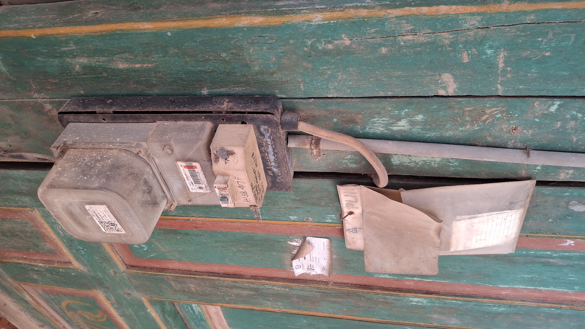 The image shows an outdoor electrical meter mounted on a weathered, painted wooden door in Pucakwangi, Indonesia. The meter is dusty and appears to be old, with a plastic cover and a metal casing. Below the meter, a beige paper envelope is attached to the door, and another piece of paper is taped to the door to the left of the meter. A curved pipe extends downwards from the meter and is attached to a vertical metal conduit. The wooden door is painted green with red and brown trim, and the paint is peeling in several places. The scene suggests a rural or older urban setting in Indonesia, likely on the exterior of a residential or commercial building.
