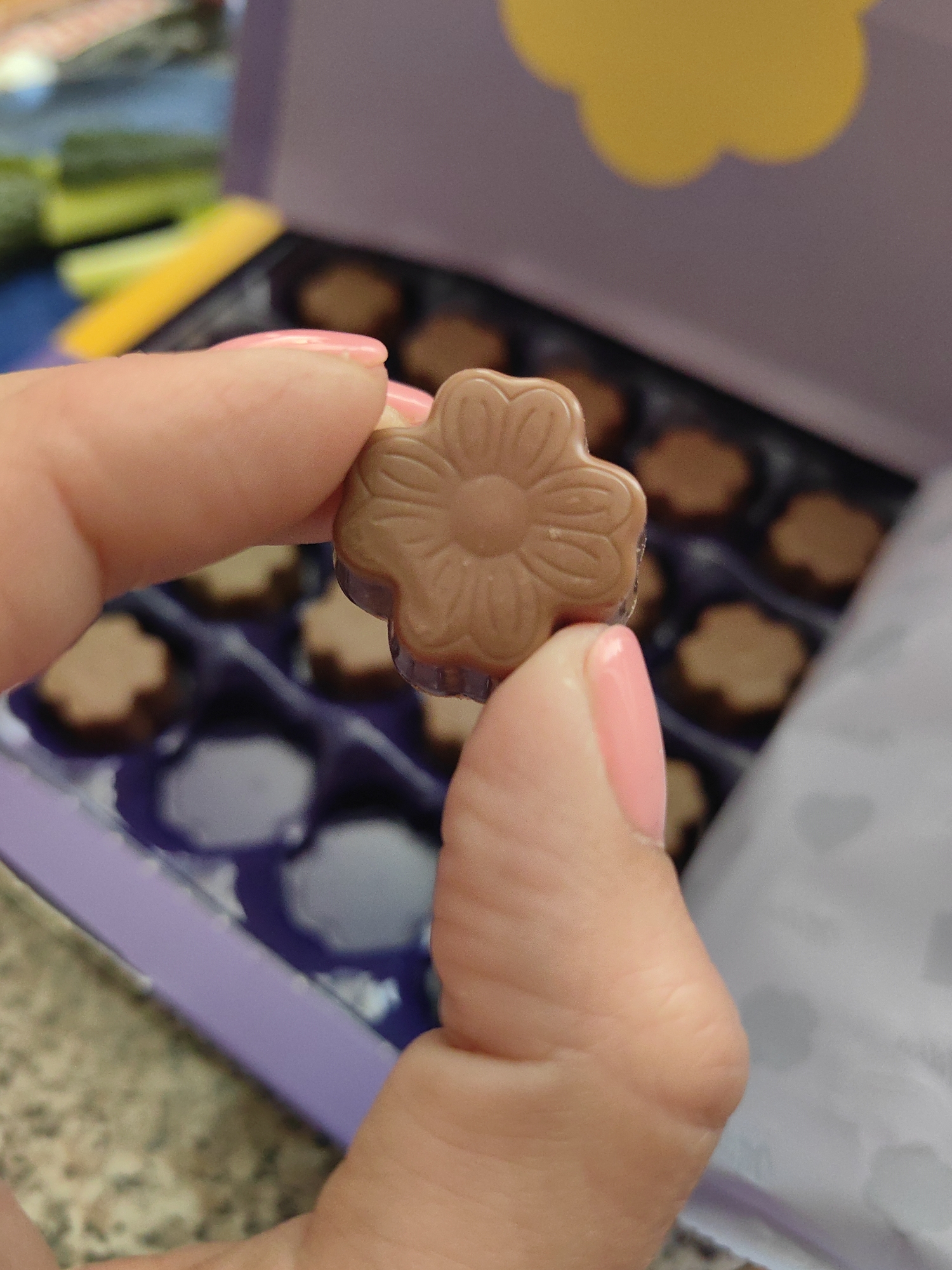 A close-up shot captures a person's hands holding a light brown, flower-shaped chocolate. The chocolate has a detailed floral design on its surface with five petals and a central circular indentation. The person's fingernails are painted a bright pink. In the blurred background, a purple box of chocolates is visible, with more chocolates arranged in a grid. A yellow object and green items are also partially visible in the background. The scene appears to be indoors, possibly in a home or a shop. No specific location in Myadzyel, Belarus can be determined from the image.