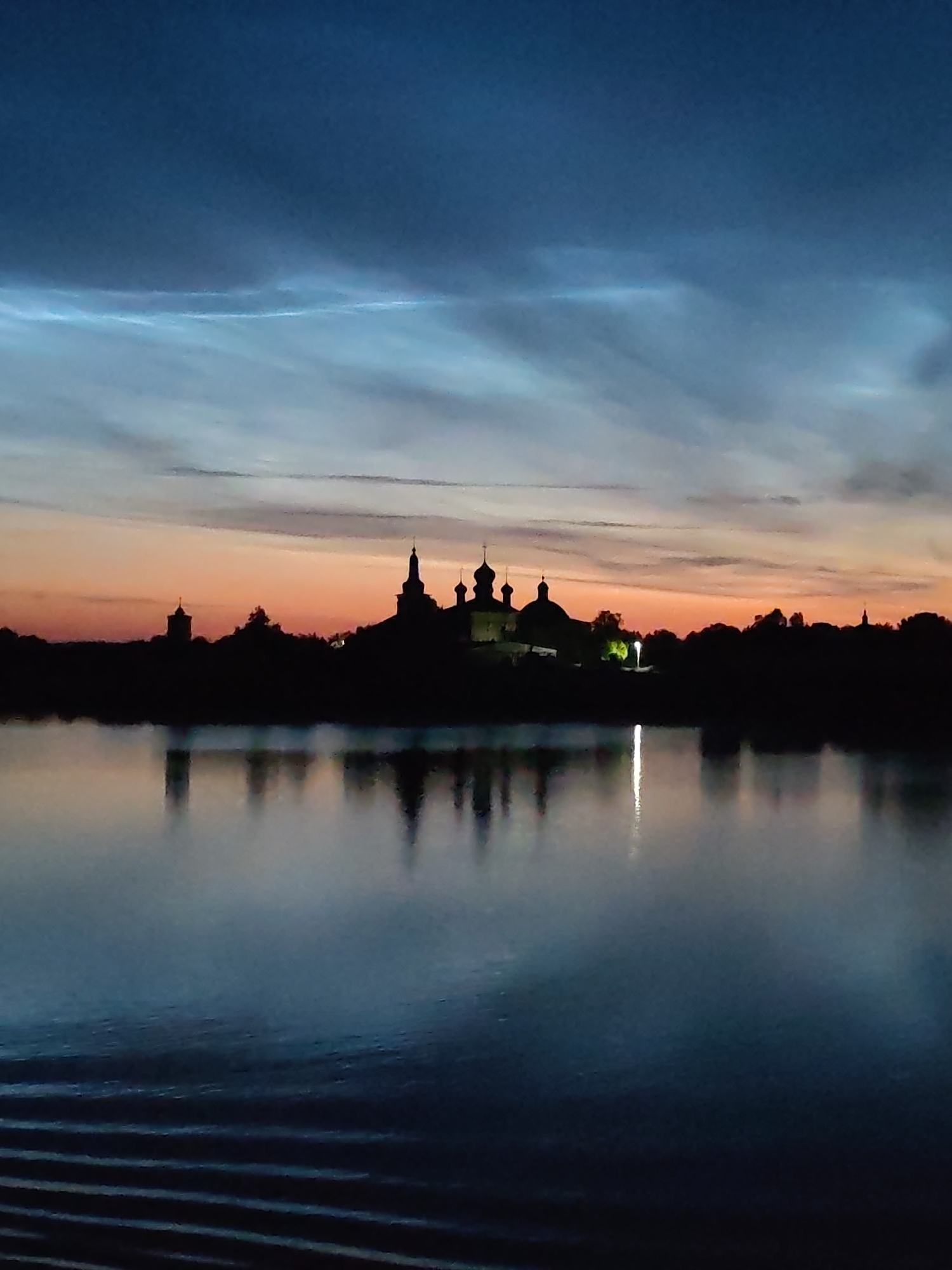 The image shows a silhouette of the Voskresensky Goritsky Monastery in Goritsy, Russia, across a body of water. The sky is a deep blue with striking noctilucent clouds, characterized by their electric blue and white wisps. The sunset colors are visible on the horizon, with shades of orange and pink blending into the darker sky. The monastery's domes and towers are clearly defined against the colorful sky, and its reflection is mirrored in the calm water. The water's surface is mostly smooth, with subtle ripples and a faint reflection of light from the monastery. The overall scene is one of tranquility and natural beauty at dusk.