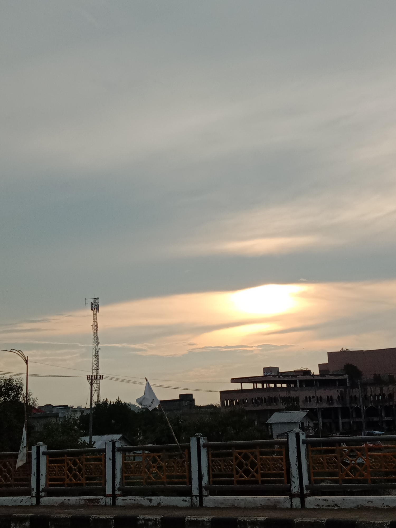 The image shows a view in Lhokseumawe, Indonesia, at what appears to be dusk. The sky is filled with clouds, with the setting sun visible as a bright area behind a layer of clouds.
In the foreground is a bridge railing painted with a combination of white, black and yellow paint. Behind the railing, there's a partially obscured view of buildings, trees, and infrastructure.
A tall telecommunications tower is visible to the left of the frame. A white flag is fixed to the railing of the bridge in front of a building. The building appears to be multi-storied and shows signs of age or disrepair.