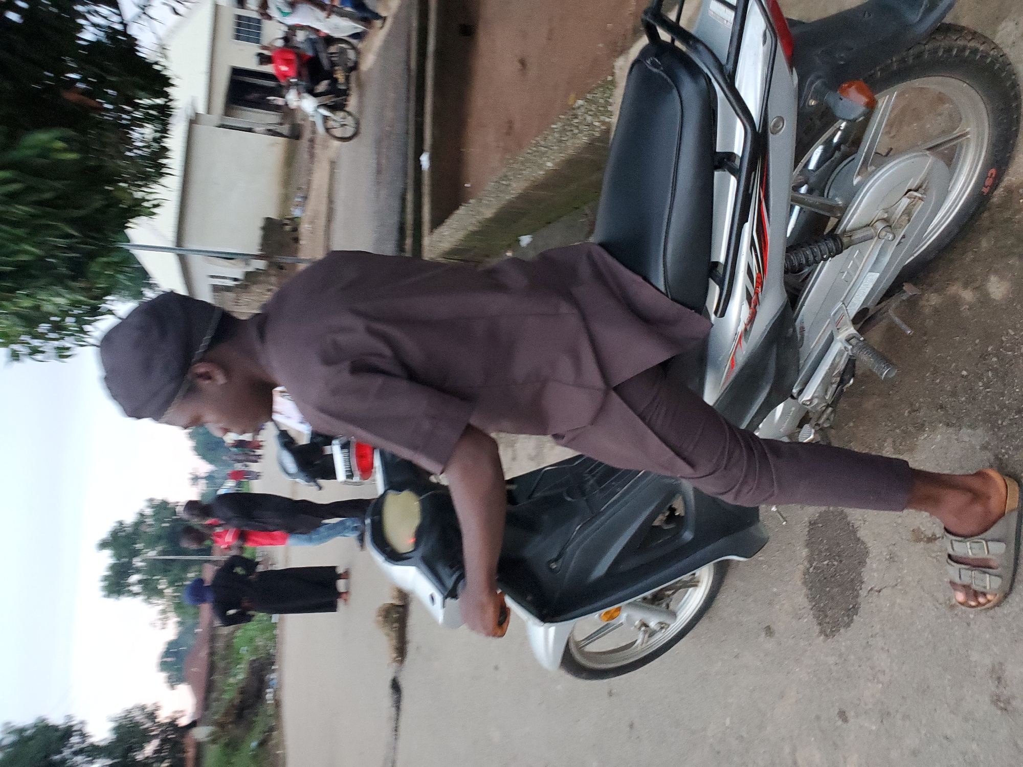 The image depicts a street scene in Mararaba, Nigeria, during daylight hours, likely late afternoon or early evening under an overcast sky.
In the foreground, a dark-skinned male, appearing to be a young adult, is seated astride a silver and black scooter-style motorbike. He wears a dark brown tunic, dark brown trousers, and a dark cap. His right hand rests on the motorbike's right handlebar. He is wearing grey slip-on sandals with two buckles. The motorbike features a black seat, silver body panels with red and black decals, a black front fairing, and a silver exhaust system. The rear tire sidewall displays "CST" in red lettering. The motorbike is positioned on a paved road.
In the midground to the left, three individuals are standing on the paved road. The leftmost person wears a dark blue head covering and a long black dress, standing with hands on their hips. Next to them is a person wearing a red t-shirt and light blue jeans. A third person, dressed in a dark top and trousers, stands facing the individual in the red t-shirt. Behind these individuals, green trees and several low-rise buildings with light-colored roofs are visible, along with an overgrown area.
In the midground to the right, another person wearing a red shirt is riding a dark-colored motorbike further down the street. A white, single-story building with a light-colored roof and dark window/door frames is present. A raised concrete drainage channel, partially filled with reddish-brown sediment and debris, runs parallel to the road on the right side.
The background shows more green foliage, trees, and additional indistinct structures, indicating an urban or semi-urban environment. Other people and motorbikes are discernible further along the street, suggesting routine activity. The paved road exhibits some wear and scattered debris.