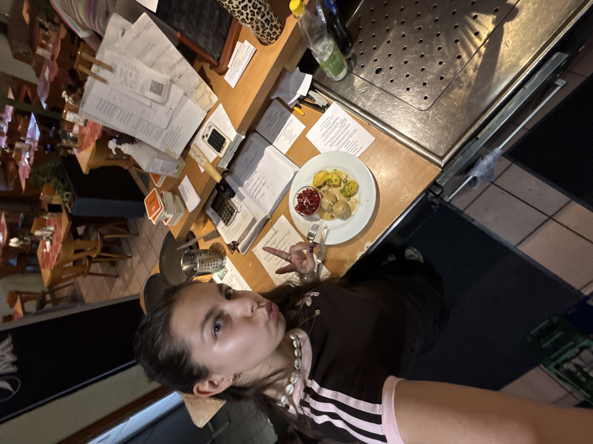A young woman is posing for a photograph with a plate of food in a restaurant. She is wearing a dark-colored Adidas shirt with white stripes and a pink collar. Her hair is brown and pulled back. She is holding up two fingers in a peace sign.
On the table in front of her is a white plate with what appears to be Swedish meatballs, mashed potatoes with gravy, and lingonberry sauce. The table is covered with papers, a calculator, and other restaurant-related items.
The background shows the interior of a restaurant with tables and chairs. The restaurant could be in Bruchköbel, Germany.