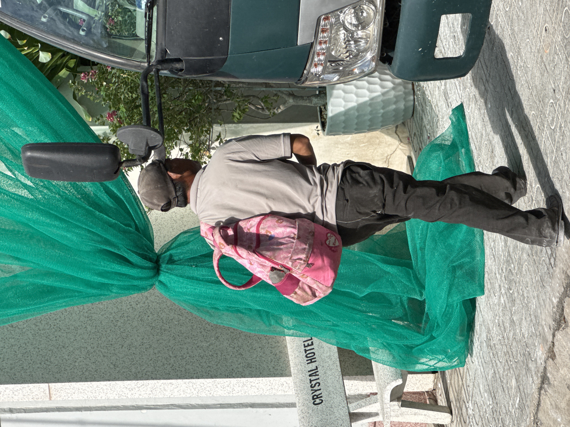 A man walks on a sidewalk near a green bus and a building with a textured facade, with a large green net draped in front of the building. The man, wearing a gray polo shirt, dark pants, and a baseball cap, carries a pink backpack with a floral pattern on his right shoulder. A white bench with the words "CRYSTAL HOTEL" is visible to the left of the man. The environment appears to be a sunny outdoor setting in a city.