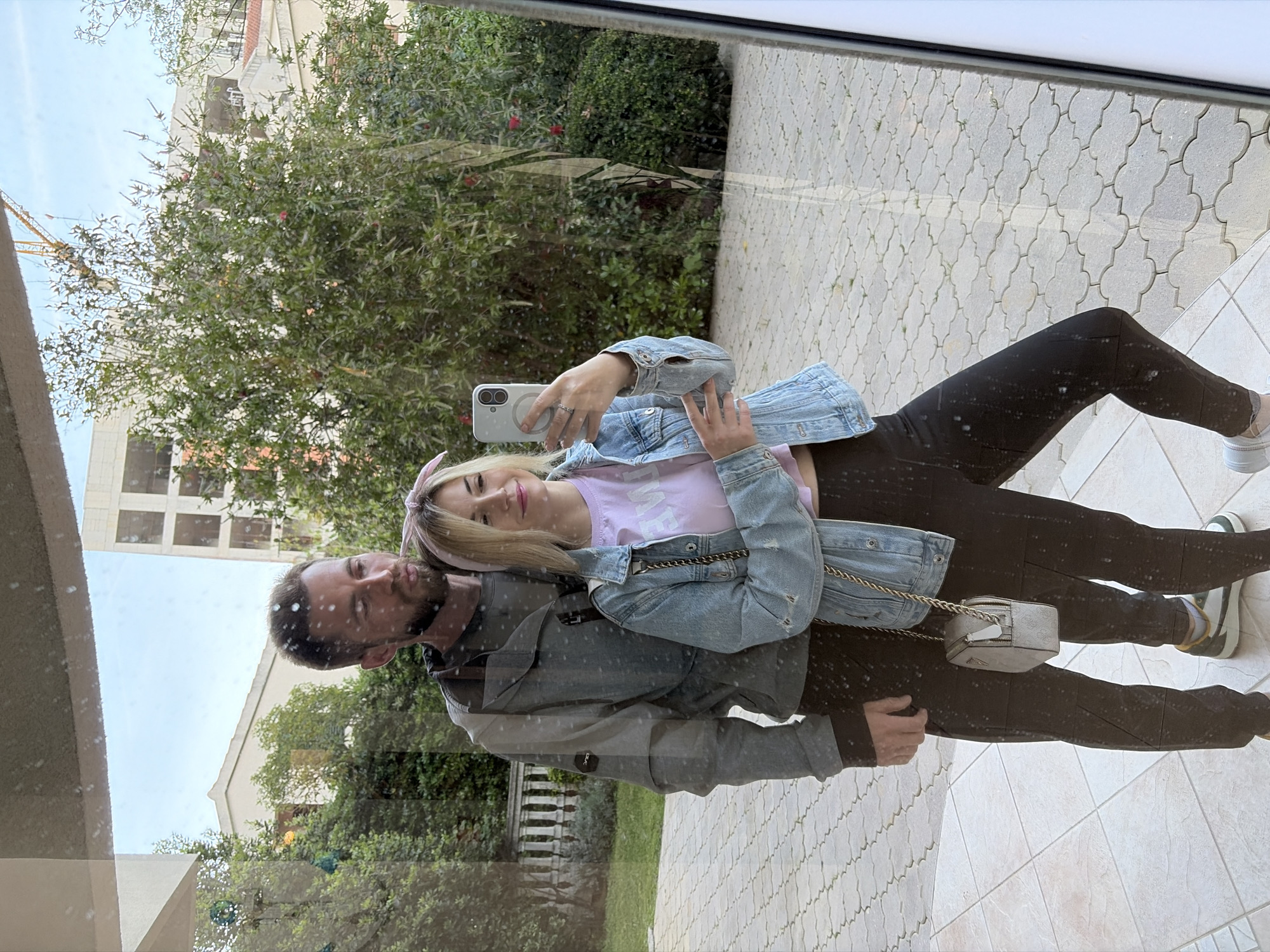 A couple is taking a selfie in front of a glass pane reflecting buildings, greenery, and a patterned pavement. The woman is holding the phone, wearing a pink shirt and denim jacket, with a pink headband. The man beside her is wearing a jacket. The location is likely in Budva, Montenegro, as indicated by the building style, plants, and pavement.