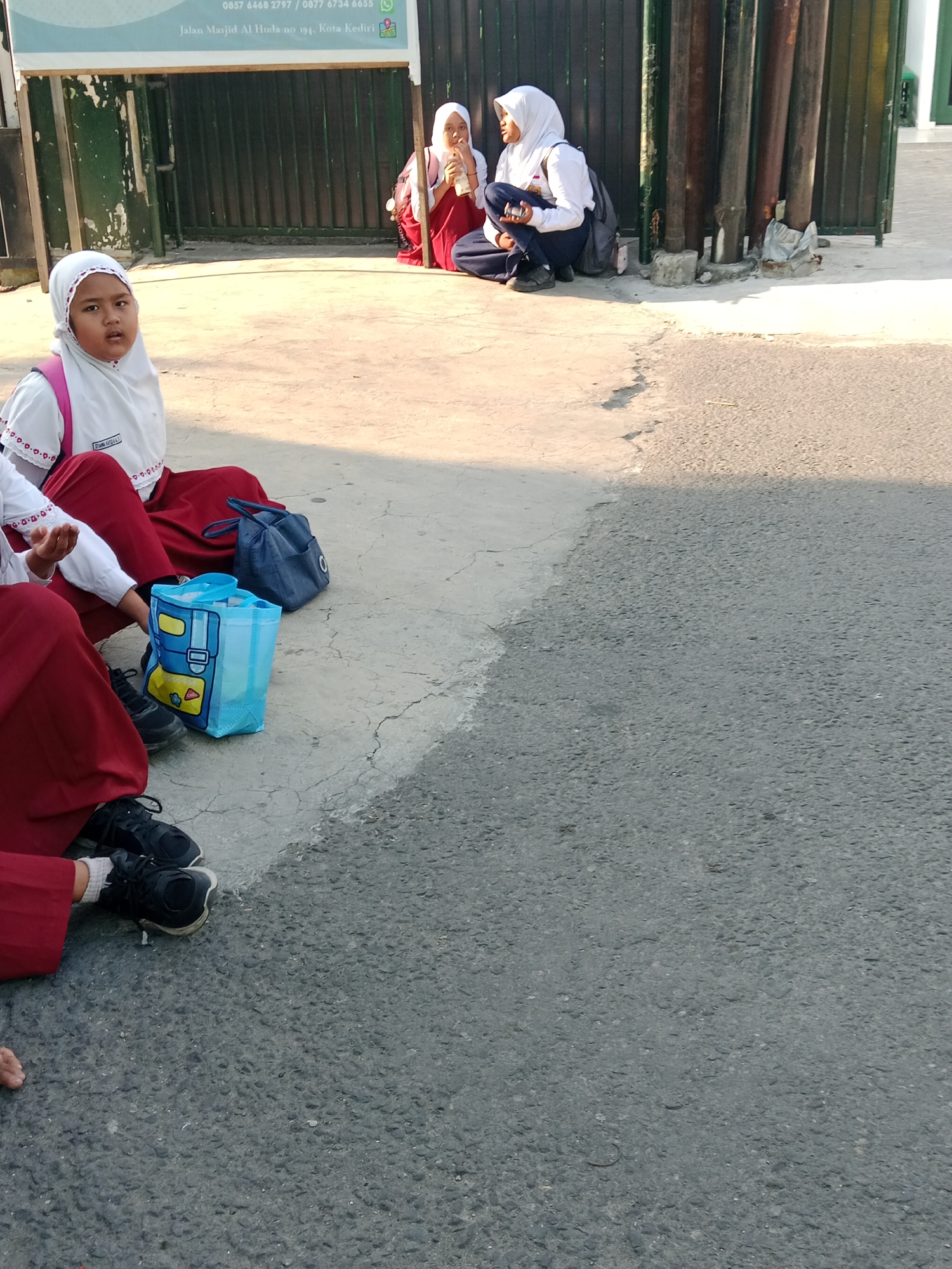 In the city of Kediri, Indonesia, three young girls are sitting on a concrete pavement next to a dark green metal fence. Two of the girls are in the foreground, wearing white hijabs and red skirts. One of them is looking directly at the camera with a pink backpack on her shoulders. The other girl's legs are visible in the foreground, wearing black shoes and white socks. Next to them are two bags, a blue one with cartoon characters and a dark blue fabric bag. Further back, another girl in a white hijab and dark blue pants is sitting on the ground, holding a drink and looking to the side. The background features a dark fence and some metal pipes. The image is taken from a slightly elevated angle.