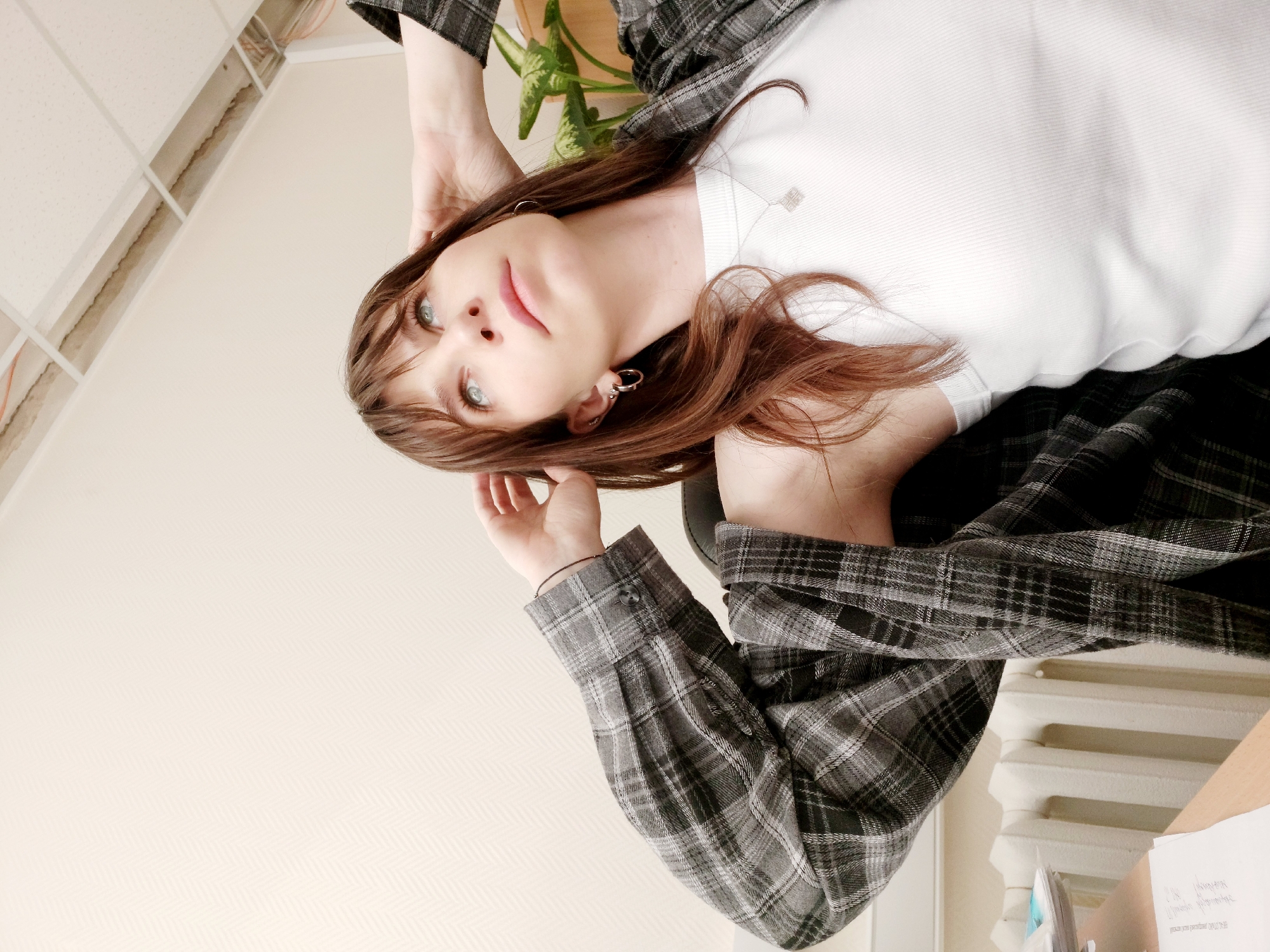 A young woman with long, brown hair exhibiting reddish undertones and striking light blue-green eyes gazes directly at the viewer from an indoor setting. She wears a white ribbed t-shirt, featuring a subtle square logo (possibly Givenchy) on its upper left chest, layered under an open dark grey and black plaid long-sleeved shirt. Her pose is relaxed, with her left hand gently touching her hair near her forehead and her right hand positioned further back on her head. She accessorizes with small hoop earrings and a delicate silver necklace with a pendant. A thin dark string is visible on her left wrist. Her expression is serene and contemplative.
The background elements suggest an office or institutional environment located in Chaikovsky, Russia. In the upper left of the frame (when oriented correctly), a suspended ceiling with white square panels is partially visible, with some sections appearing unfinished or removed, exposing wiring and structural components above. A large, leafy green potted plant stands behind her. The walls are a light, neutral tone, with a subtle diagonal pattern visible on the wall to the lower right. A white radiator is partially visible in the lower right corner, adjacent to a surface that holds several papers, on which some faint and illegible Cyrillic text can be discerned. The room is brightly and evenly lit, characteristic of indoor daylight or artificial lighting.