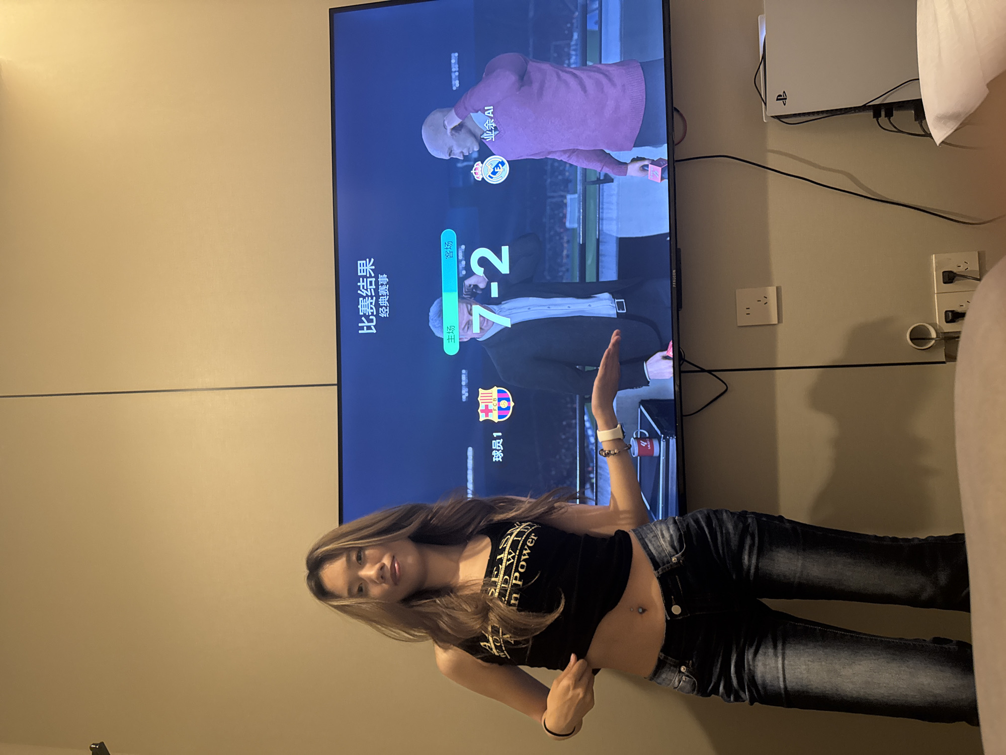 A waist-up photograph shows a young woman in a black tank top and faded black jeans in an indoor setting. The woman is facing and slightly angled toward the viewer. She has light-brown hair and her midriff is exposed. Behind her is a tan wall with a large television mounted on it. The TV screen displays a video game interface. A PlayStation console is above the television on the wall. Wall sockets are present to the right of the television. The language on the screen is Chinese. It says "比赛结果 经典赛事" translating to "Match results, Classic match". The score on the screen is 7-2. The logo for FC Barcelona is also visible on the screen. The photo was most likely taken indoors in a room that is at the city of Liangjiang, China.