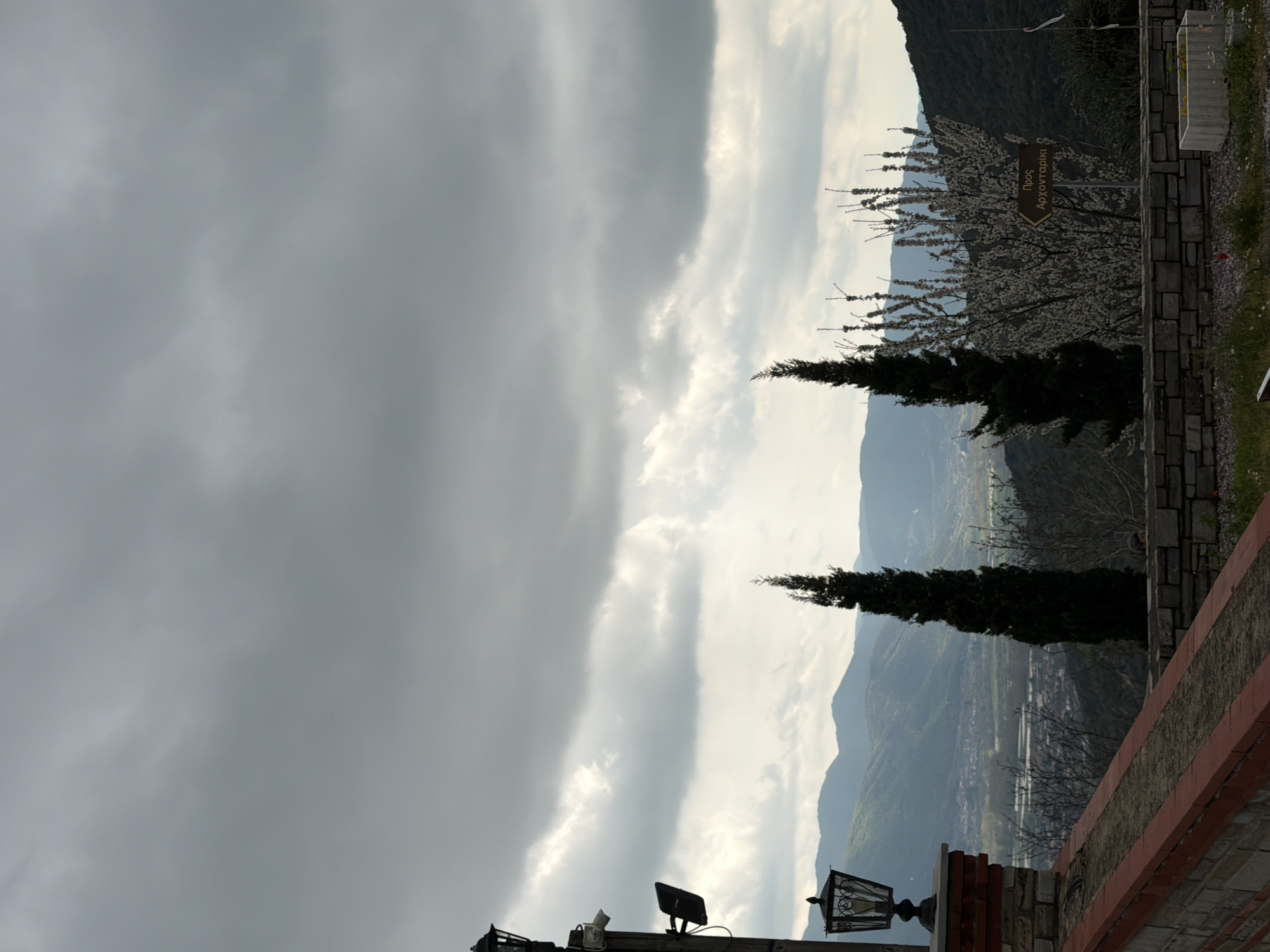 The image shows a landscape in Chrysokastro, Greece, under an overcast sky. The view includes mountains in the background and two tall, slender cypress trees in the midground. A stone wall with a red-painted top edge is visible in the foreground, along with a vintage-style street lamp mounted on a brick and stone structure. A signpost with Greek text is partially visible amidst blossoming trees. The overall environment appears to be an elevated viewpoint overlooking the valley.