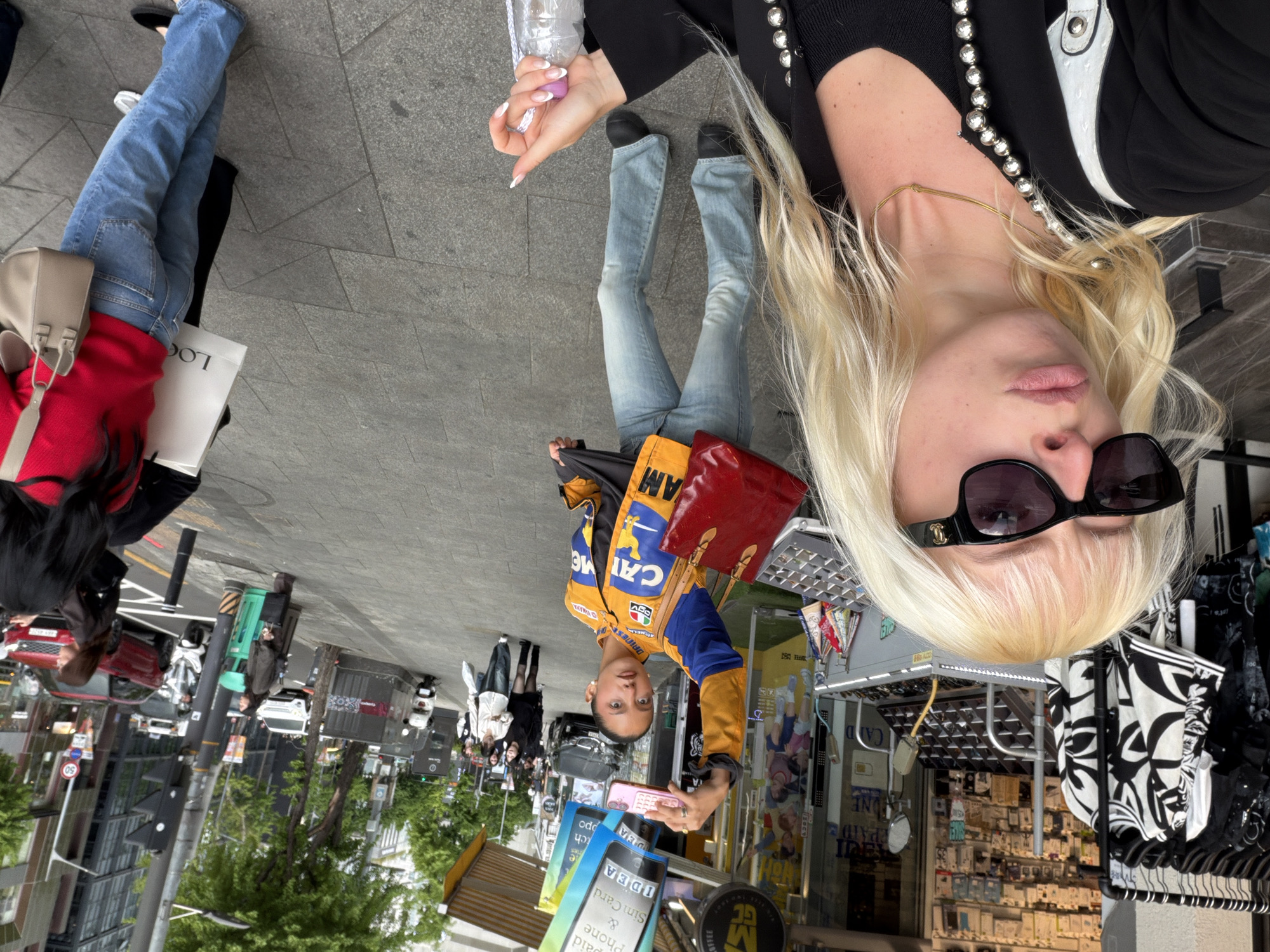 This rotated daytime image captures a vibrant street scene in Seoul, South Korea, filled with pedestrians and urban details. The perspective is tilted 90 degrees clockwise.
In the foreground, a blonde woman wearing black sunglasses with a visible Chanel logo, a black top adorned with beaded embellishments, and a gold necklace is prominent. She holds a plastic water bottle, and her long, light-colored hair cascades downwards.
Mid-ground to the left, another individual is seen wearing a distinctive yellow, blue, and black racing-style jacket with "CAI ME AM" text, light-wash jeans, and black shoes. They carry a red handbag and hold a pink smartphone, looking directly towards the viewer. Further left, another person wears a red top and blue jeans, carrying a light beige handbag and a white shopping bag with the partially visible word "LOG".
The background reveals a bustling city environment with other pedestrians, passing cars (including a red SUV), motorcycles or scooters, and various commercial establishments. Streetlights, trees, and buildings line the street.
Visible text on signs and objects includes "LOG" on a shopping bag, "CAI ME AM" and "ADD" on a jacket, and "DAINESE VINARA" and "e" also on a jacket, possibly indicating brands. Street signage shows "50" (likely a speed limit). Shop advertisements and signs display "IDEA", "Sim Card", "Phone", "Paid", "ONE", "OFFEE", and "GC", indicating businesses offering telecommunication services and coffee. The overall scene depicts casual daily life and movement within an urban setting.