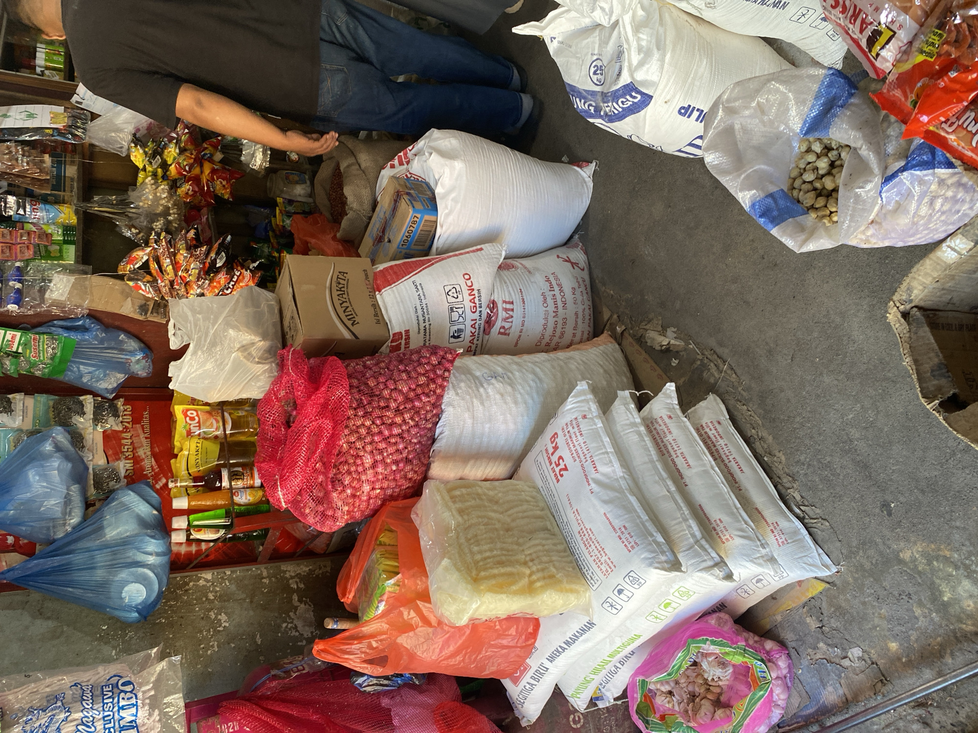 The image depicts a cluttered storefront, possibly a small market or convenience store. A person wearing a black t-shirt and blue jeans stands on the right, facing away from the camera, with their right arm bent and resting on a stack of items. The store is filled with various goods, including bags of rice or flour, sacks of produce like onions, and smaller packaged items such as snacks and bottled liquids. Several plastic bags, some filled and others empty, are visible, along with cardboard boxes and what appear to be scales or weighing equipment. The flooring is uneven and appears to be concrete with some debris. The overall impression is of a busy, perhaps slightly disorganized, retail environment.