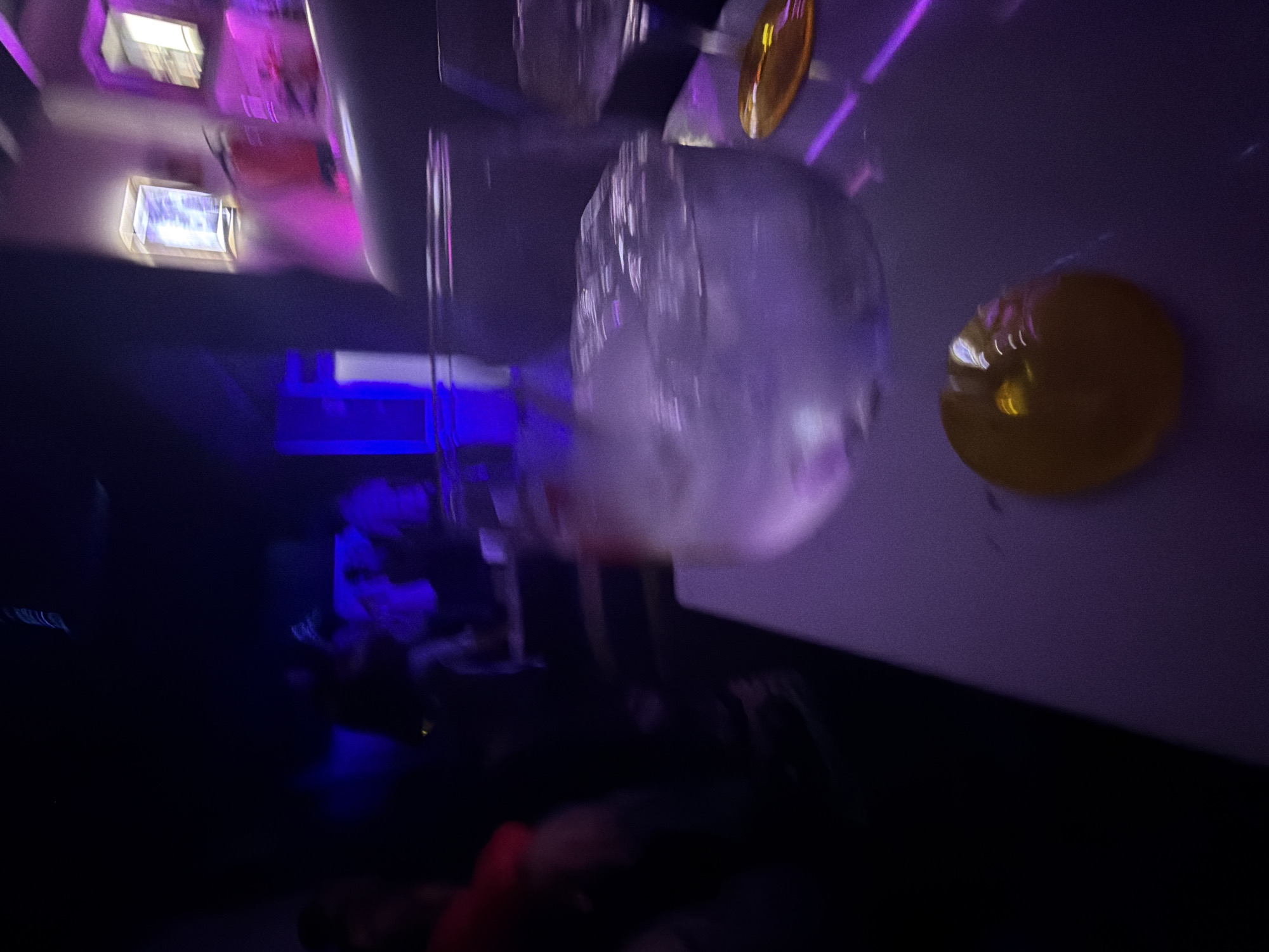 The image appears to be taken inside a venue, possibly a club or bar, in Vila Real, Portugal, judging by the interior design and lighting. The photo is blurry, suggesting motion.
**Subjects and Objects:** A translucent object, possibly a plastic bag or a balloon, is suspended from the ceiling. There are illuminated fixtures, including ceiling lights and a yellow spherical light fixture. In the background, indistinct figures of people are visible, appearing to be seated or standing near blue lighting.
**Environment:** The environment is dimly lit with primarily blue and purple lighting, typical of a nightlife setting. The ceiling appears white.
**People:** People are present in the background, though their actions and interactions are not discernible due to the blur and darkness.