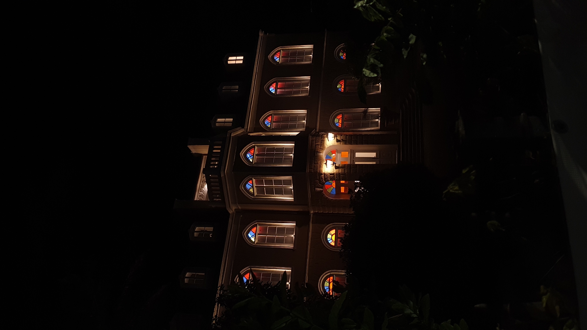 The image shows a multi-story building at night, likely located in Bar, Montenegro based on the user prompt. The building has a distinctive architectural style, featuring arched windows with colorful stained glass details in red, blue, and yellow hues. Some windows are brightly illuminated from within. The building appears to be made of a light-colored material. A staircase is visible on the left side of the building. The surrounding environment is dark, with foliage partially obscuring the lower portion of the building and a dark expanse in the background. A light-colored vertical structure or element is visible on the right side of the image, likely a building or architectural feature.
