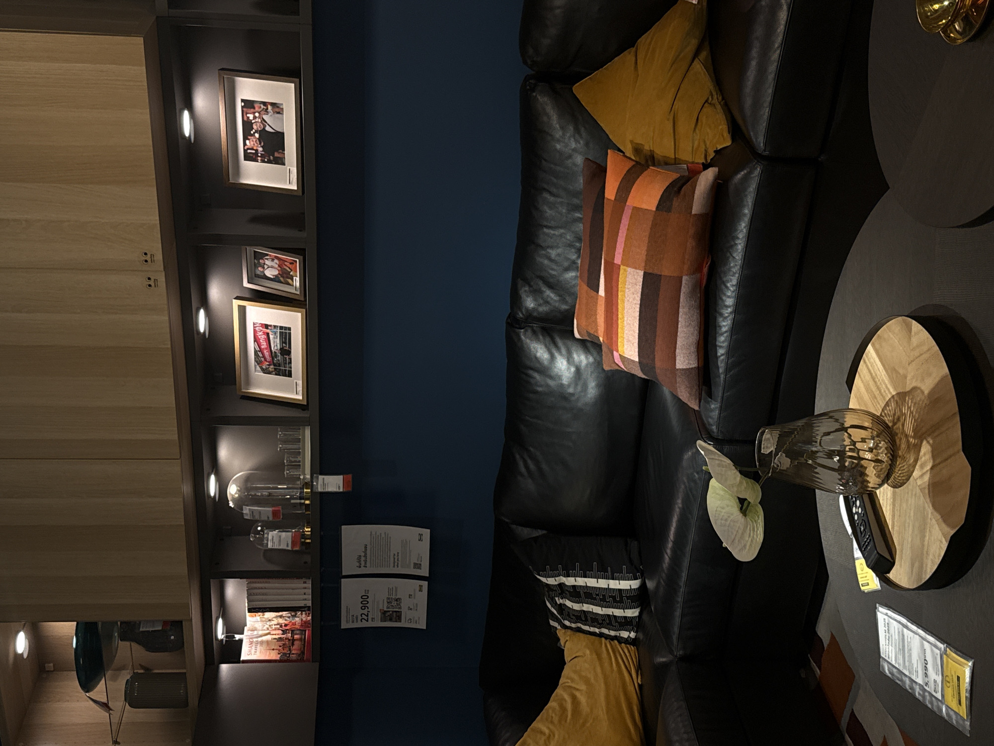 The image depicts a cozy living room setting with a dark blue wall as the backdrop. A plush black leather sectional sofa dominates the scene, adorned with several decorative pillows. One pillow features a geometric pattern in shades of orange, brown, and pink, while another is a solid mustard yellow. A white flower rests on the sofa, adding a touch of elegance.
To the left of the sofa is a modern shelving unit with integrated lighting. Several framed photographs are displayed on the shelves, capturing moments with people. There are also glass decorative items and books on the shelves.
In the foreground, a dark round coffee table is visible with a wooden circular inlay. On the table, a glass vase with a textured pattern holds a white flower. A remote control lies next to the vase. A price tag with Thai text and the amount "22,900" is attached to an item on the shelf to the left, and another price tag with "5,990" is on the table.
The scene suggests a comfortable and stylish indoor environment. The lighting is soft, indicating it could be evening or a dimly lit day. The presence of price tags suggests the setting might be a furniture showroom or a display within a store, possibly in Bangkok, Thailand, given the Thai text on the tags. The overall mood is one of relaxation and home comfort.