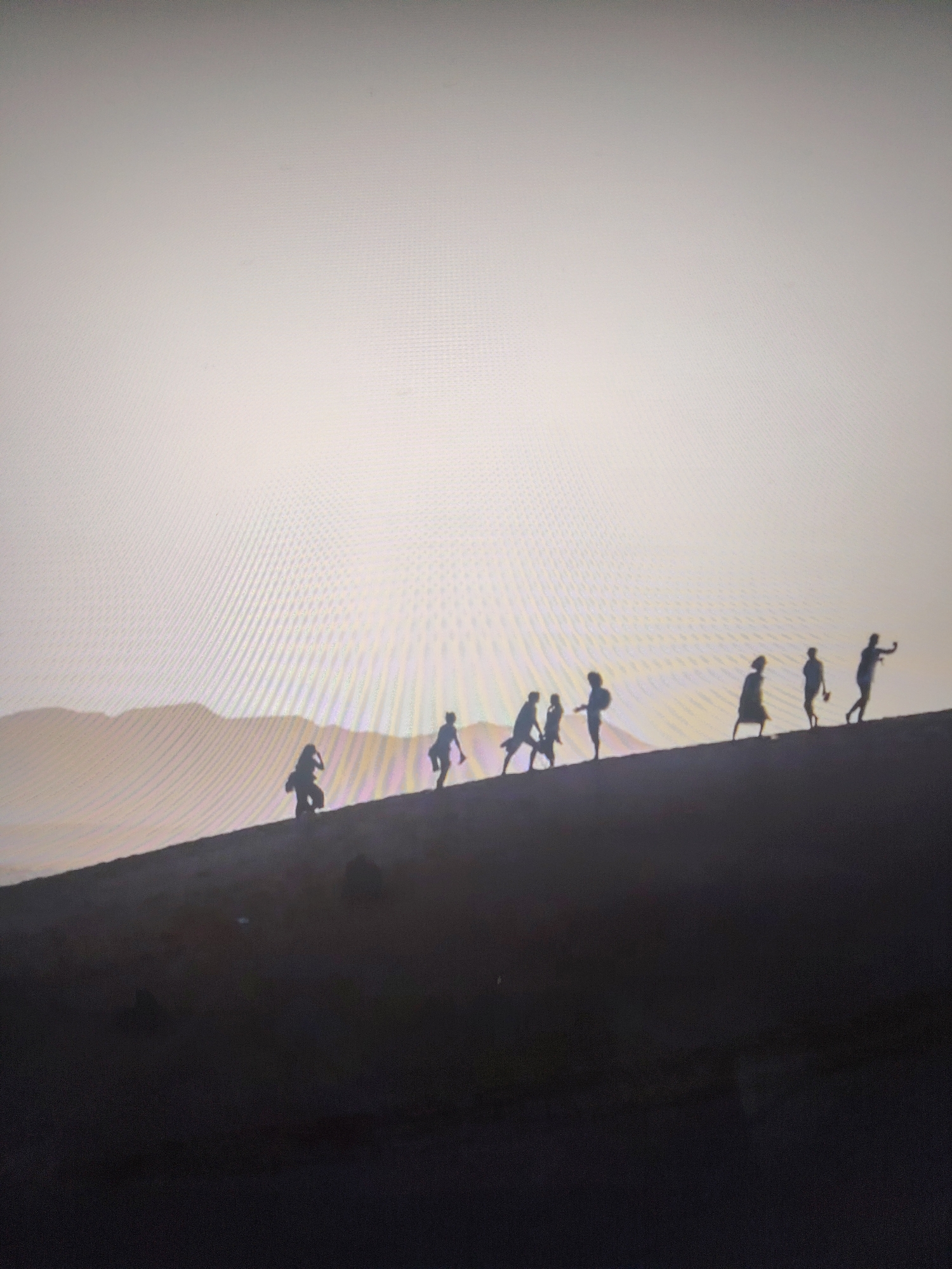 A line of silhouetted figures is visible atop a ridge against a light-colored sky. The ridge appears to be a sand dune, with further landmasses visible in the distance behind it. The location is Unknown, Unknown.