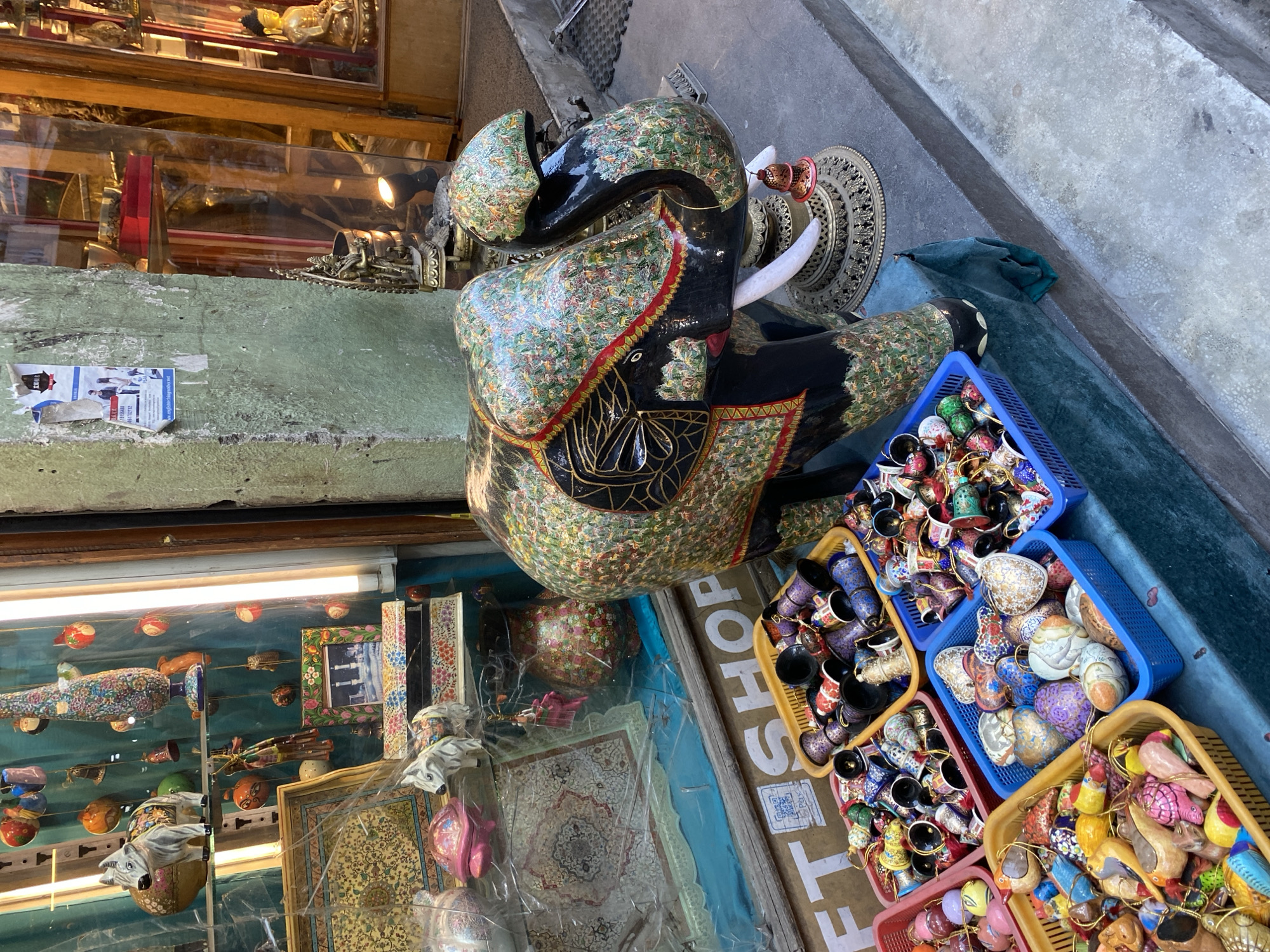 The image captures a close-up of a large, ornate, decorative elephant statue placed on a stone step outside a shop. The elephant is adorned with intricate, colorful patterns. In front of the elephant and to the left, several plastic baskets are filled with small, decorative items, possibly ornaments or handicrafts, including bells, birds, and egg-shaped objects. Some of these are also visible through a shop window behind the elephant. A partially visible sign on the shop window reads "GIFT SHOP." The shop appears to be located in a marketplace or commercial area within Kathmandu Metropolitan City, Nepal, given the style of the goods and the general aesthetic. The surrounding environment includes a textured stone wall to the left, a glass shopfront reflecting various items, and the concrete pavement. No people are visible in the frame.