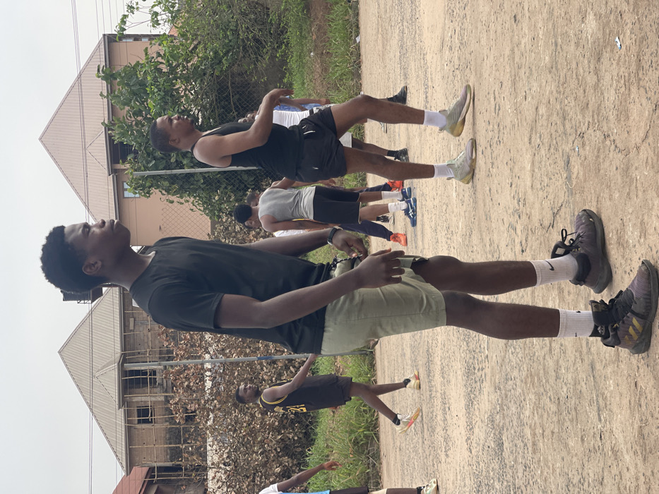 The scene depicts an outdoor gathering of several young adult males on an unpaved, light brown dirt or gravel surface in Umunekwu, Nigeria, under an overcast sky.
In the immediate foreground, a tall, slender Black male stands in profile, facing right. He wears a black short-sleeved t-shirt, light olive green shorts, white socks with a black Nike logo, and dark grey and gold Adidas high-top sneakers. His left arm is slightly bent, and he has a dark band or watch on his left wrist. Adjacent to him on the right, another Black male of medium build stands in profile, also facing right, wearing a black tank top, black shorts, white socks, and light green/yellowish athletic shoes.
Behind these two figures, several other Black males are visible across the mid-ground. To the left, a male in a black basketball jersey with a yellow "24" on the back and black shorts walks right, looking back over his left shoulder. Further back and to the right, a male in a grey tank top and black shorts is seen from behind, facing away. Other partially obscured individuals wear various athletic attire, including t-shirts, tank tops, shorts, and brightly colored athletic shoes (e.g., orange, blue, white).
The background features residential structures and vegetation. To the left, a two-story building under construction, showing unpainted brickwork and visible scaffolding, has a brown corrugated metal roof. Adjacent to it on the right, a finished two-story building with a light brown facade also features a brown corrugated metal roof. Dense green foliage and dry brown bushes are present behind and between the buildings, and a wire mesh fence is partially visible on the far left. The ground shows some minor scattered debris. The overall impression is a casual assembly, likely related to sports or exercise, in a semi-urban or residential area.