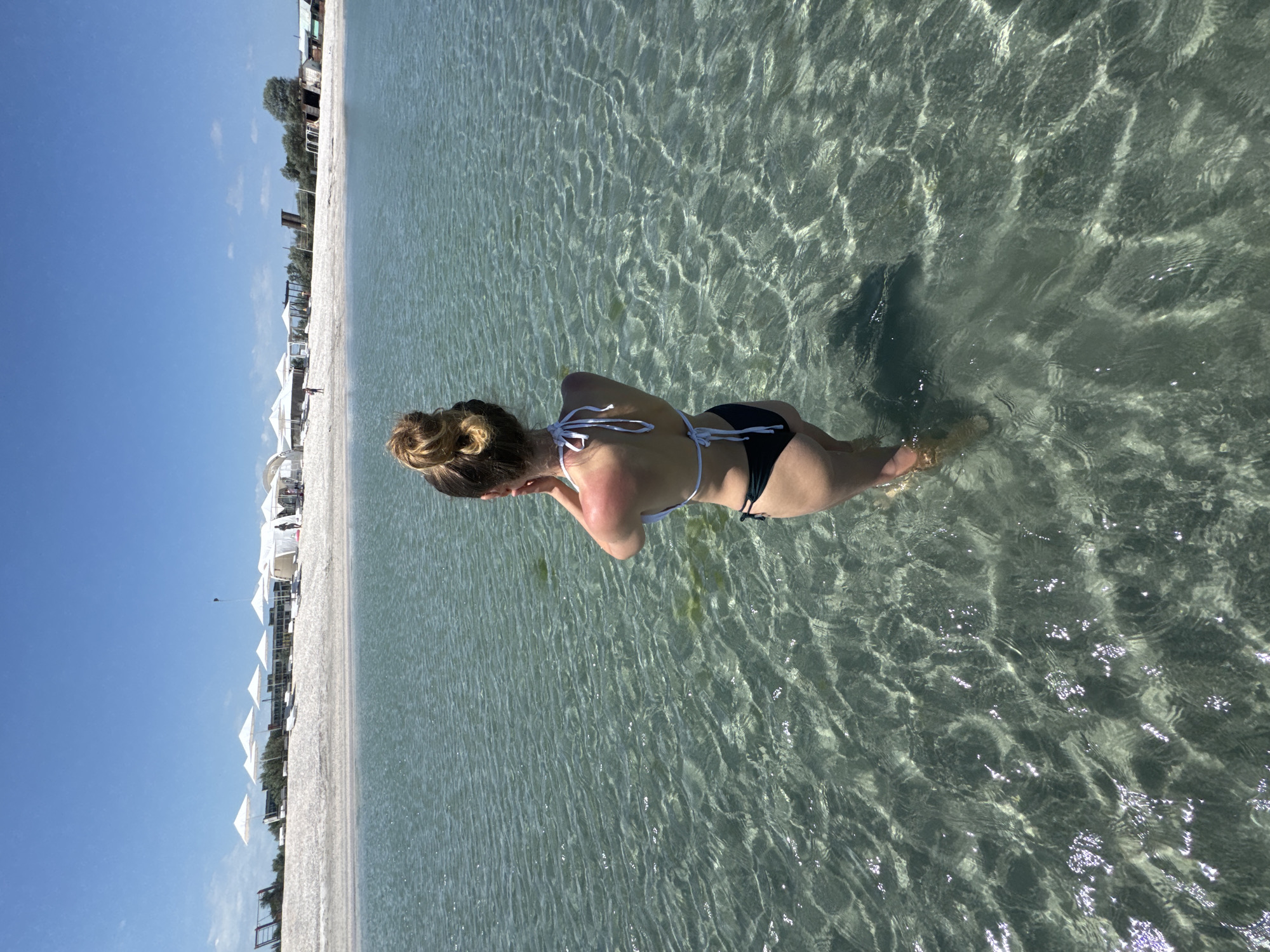 A woman in a bikini stands in the clear, shallow water of a beach in Мысовское сельское поселение, Ukraine. The beach, lined with white umbrellas, is visible on the left side of the frame under a blue sky. The water reflects sunlight.