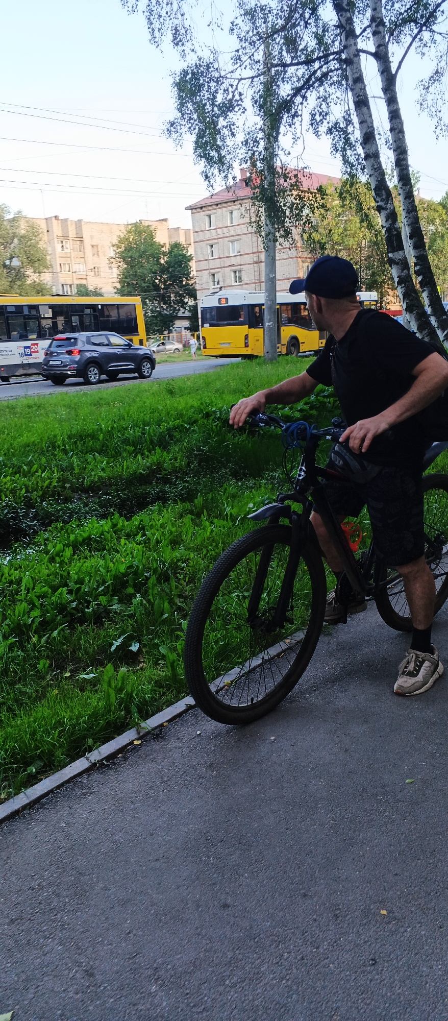 A man in a black t-shirt, black shorts, and a navy blue baseball cap is standing next to a black bicycle. He is facing away from the camera, looking towards the left of the frame. The bicycle has a blue cable lock around the handlebars. The man's right hand is on the handlebar. In the background, there is a street with two yellow buses, one with the number 21 visible. A dark gray SUV is driving on the street. There are buildings and trees visible behind the street. The scene appears to be in an urban environment, likely a residential area, possibly in Izhevsk, Russia, given the architectural style of the buildings and the type of buses. The ground is paved, with a grassy area bordering the pavement. The lighting suggests it is daytime, possibly late afternoon or early morning.
