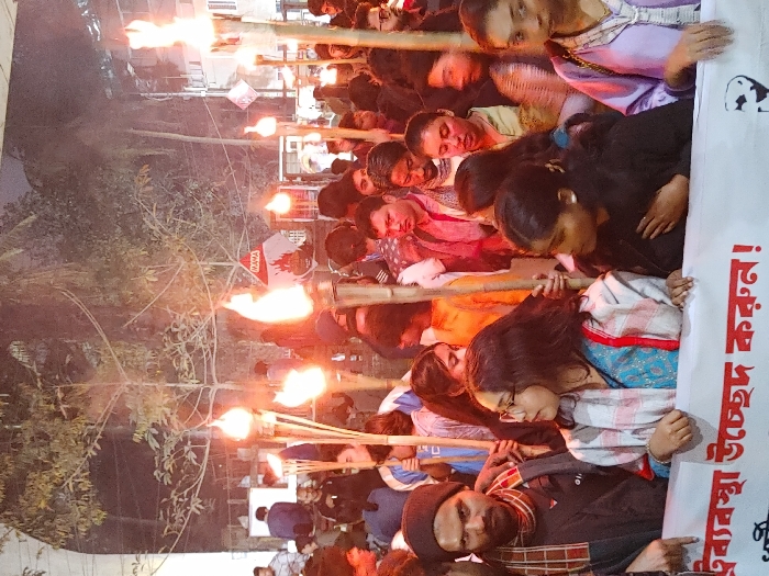 The image depicts a night-time torch procession in Dhaka, Bangladesh, likely situated within the Dhaka University campus or the vicinity of the Shahbag intersection. A dense crowd of individuals, predominantly young adults, moves or stands in close proximity. Numerous participants are holding ignited bamboo torches (mashals), which serve as the primary light source, casting strong orange and yellow hues and creating a smoky atmospheric effect.
In the immediate foreground, a white banner displaying red Bengali text is partially visible, reading "ঐ ব্যবস্থা উচ্ছেদ করুন !" (Abolish that system!). Prominent individuals include a man on the far left wearing a black beanie and a multi-patterned, possibly checkered, scarf over a dark top, looking towards the viewer. To his right, a woman with glasses wears a blue garment under a patterned white and red shawl, looking towards her right. Further to the right, another woman in a dark top places her left hand on her chest.
In the background, a white sign featuring the red word "MAMA" and an associated red graphical emblem is visible. Bare-branched trees and faint urban structures are discernible against the dark sky. The overall scene conveys an organized public protest or demonstration.