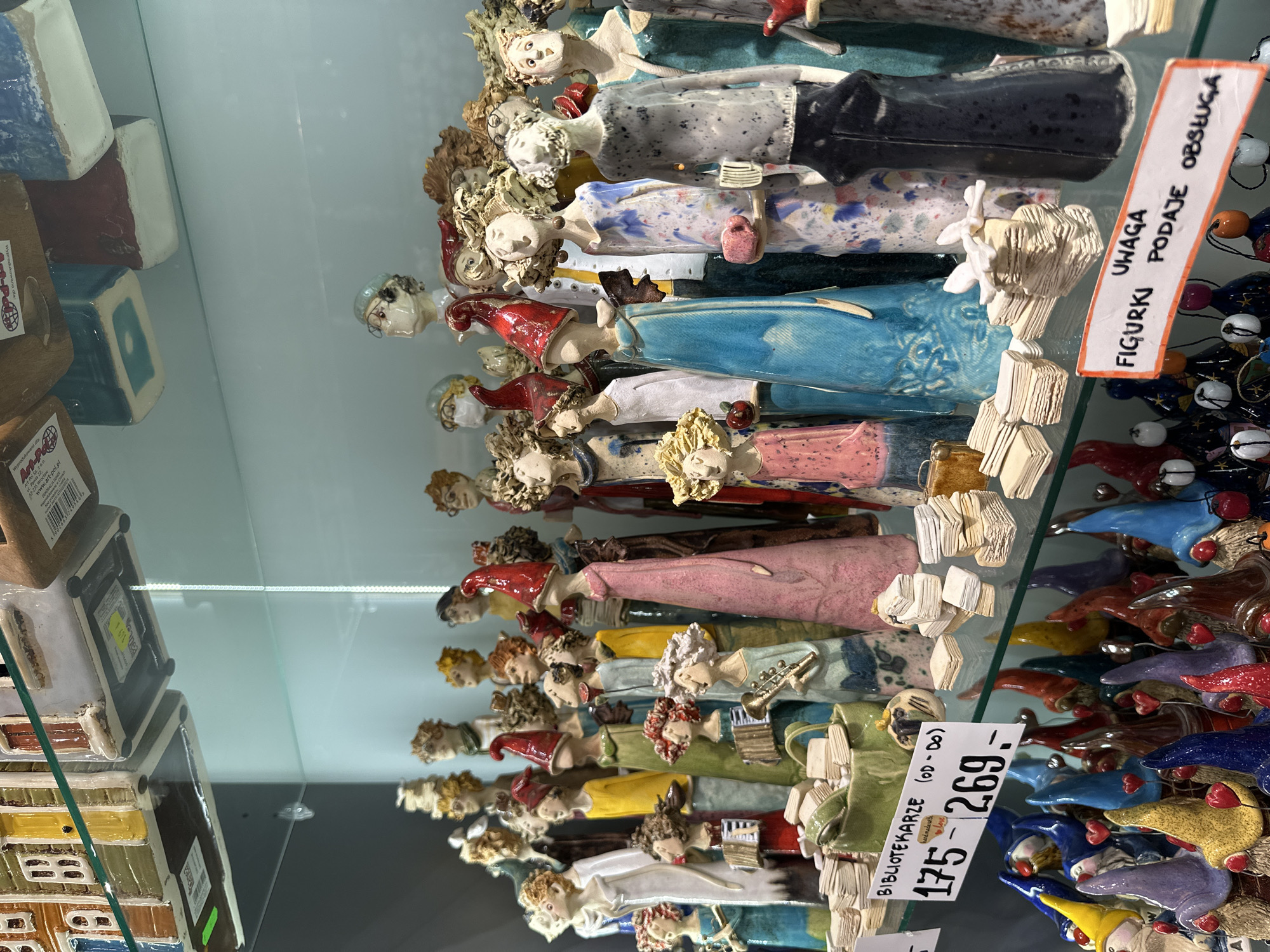 The image shows a display case filled with rows of handcrafted ceramic figurines. Each figurine appears to represent a different character, possibly related to librarians ("BIBLIOTEKARZE" is visible on a price tag). The figurines are individually painted and decorated, with varying colors and details. A sign reads "FIGURKI PODAJE OBSŁUGA", indicating that the figures are handled by staff. The photograph was taken in Wrocław, Poland.